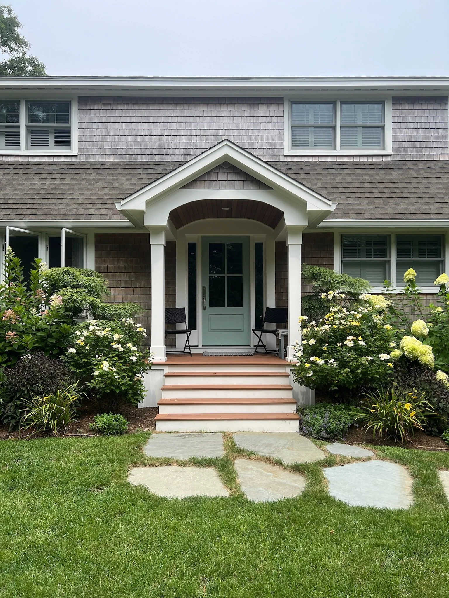 Front view of a house with a small porch, white steps, black chairs, and surrounded by green bushes and flowering plants.
