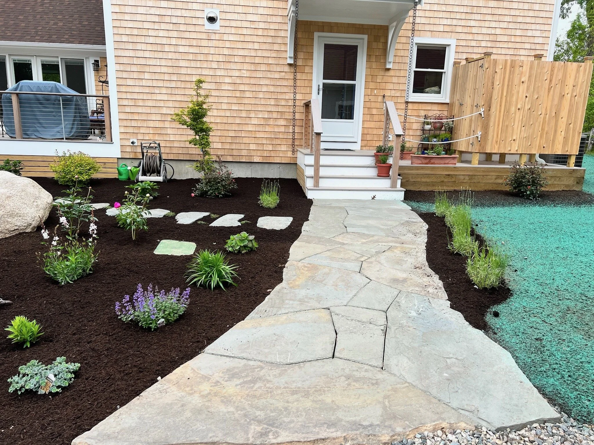 A backyard landscaping scene with a stone pathway leading to a house porch, surrounded by freshly planted flowers and shrubs, with a wooden deck on the right and a patio area on the left.