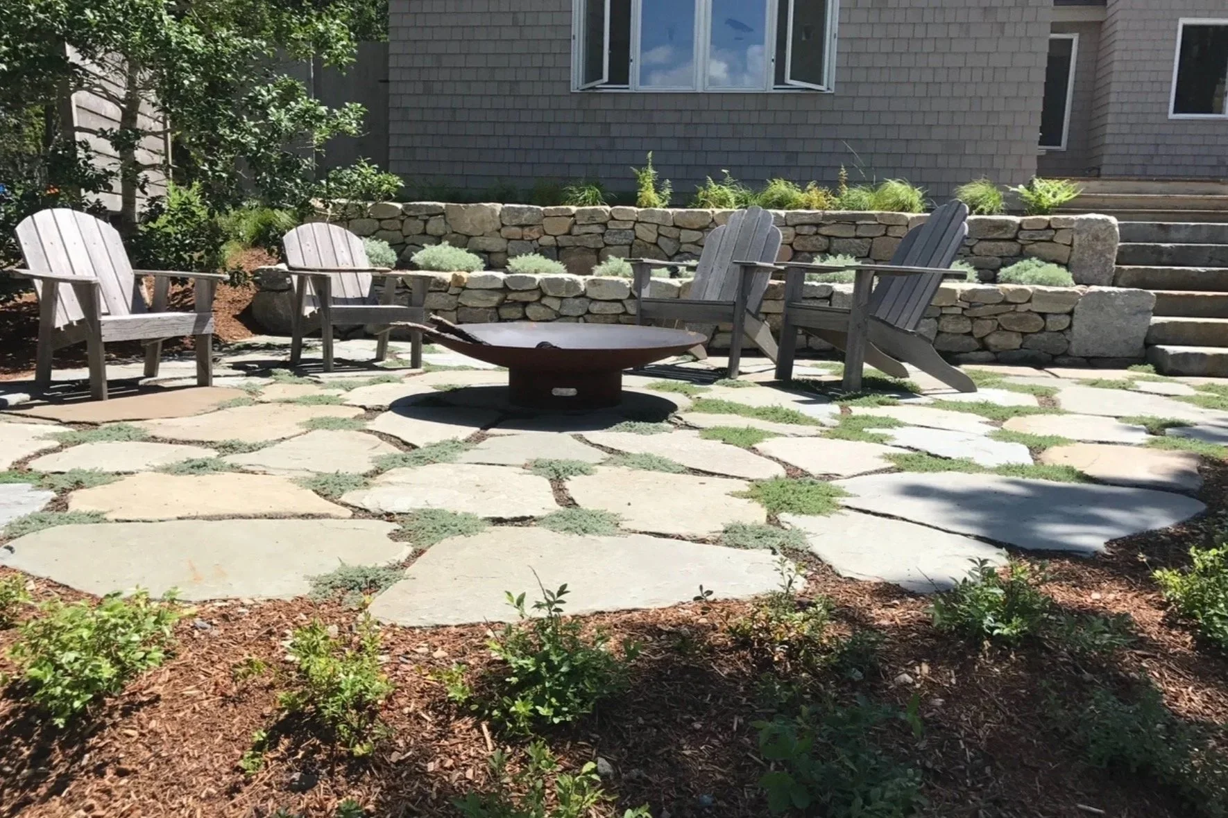A landscaped outdoor patio with large stone pavers, wooden Adirondack chairs, a metal fire pit, and stone retaining walls with plants.