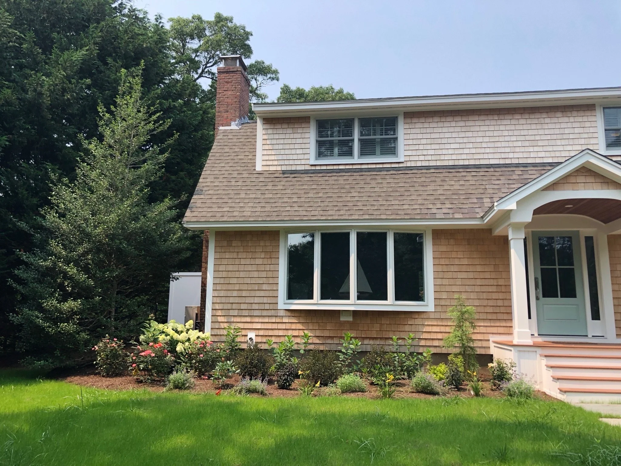 A two-story house with beige shingle siding, a gable roof, a brick chimney, and a front porch with steps. The house has large windows and a well-maintained front yard with grass, flowers, and bushes. There are tall trees in the background.