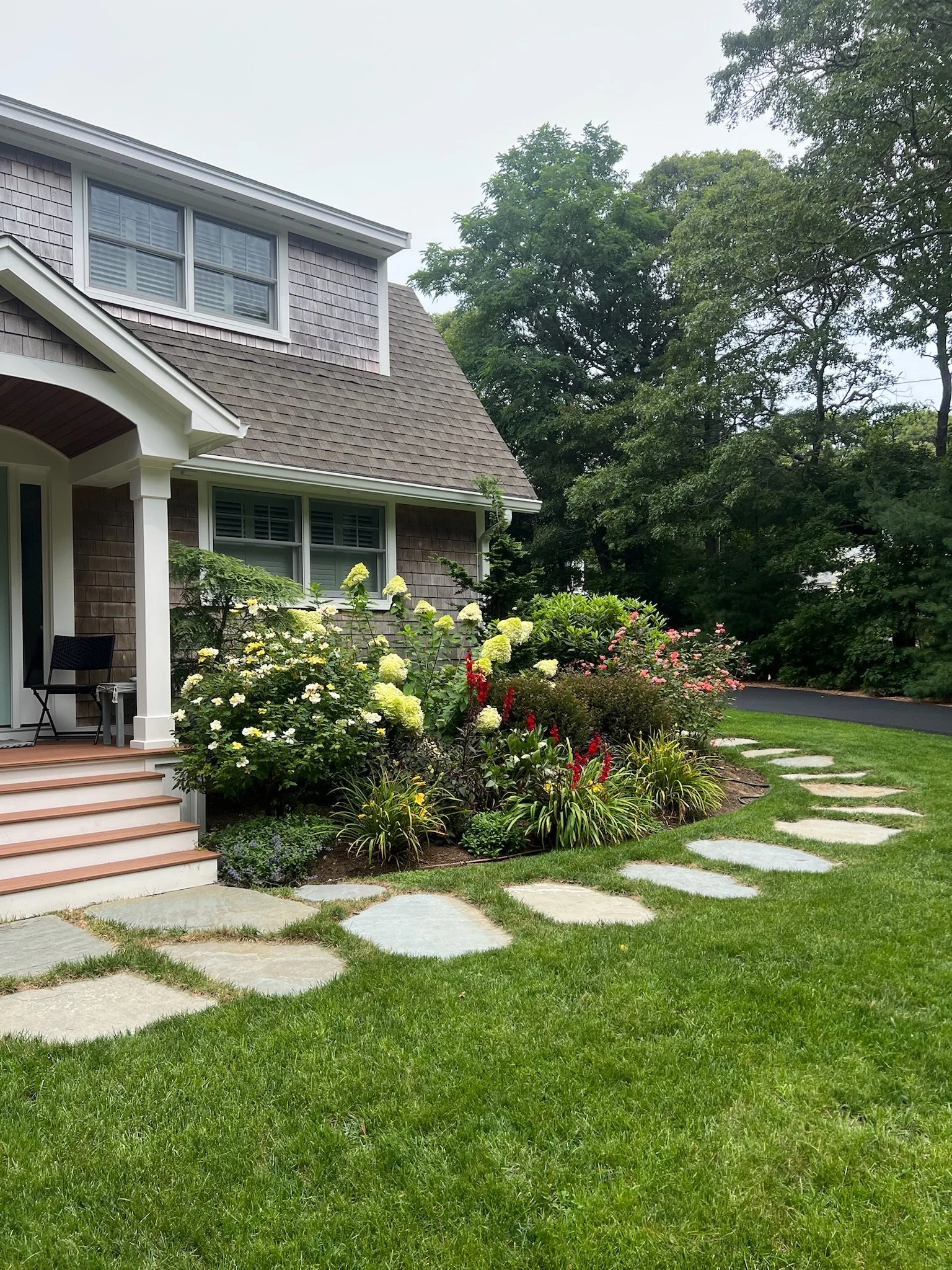 Front yard of a house with stairs, garden, and stone pathway, surrounded by green trees.