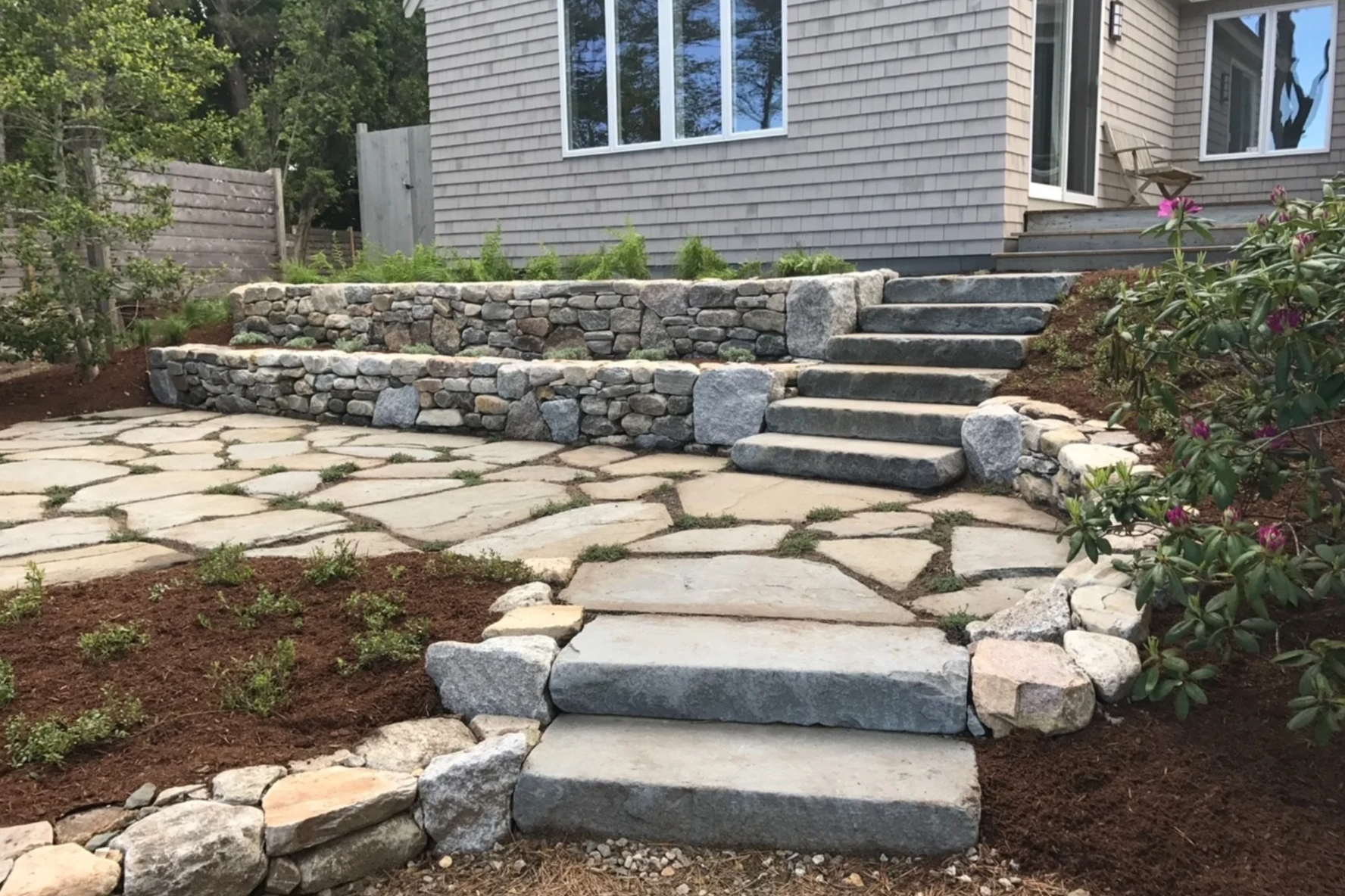 Stone steps leading up to a house with a landscaped yard featuring flagstone and stone wall retaining beds.