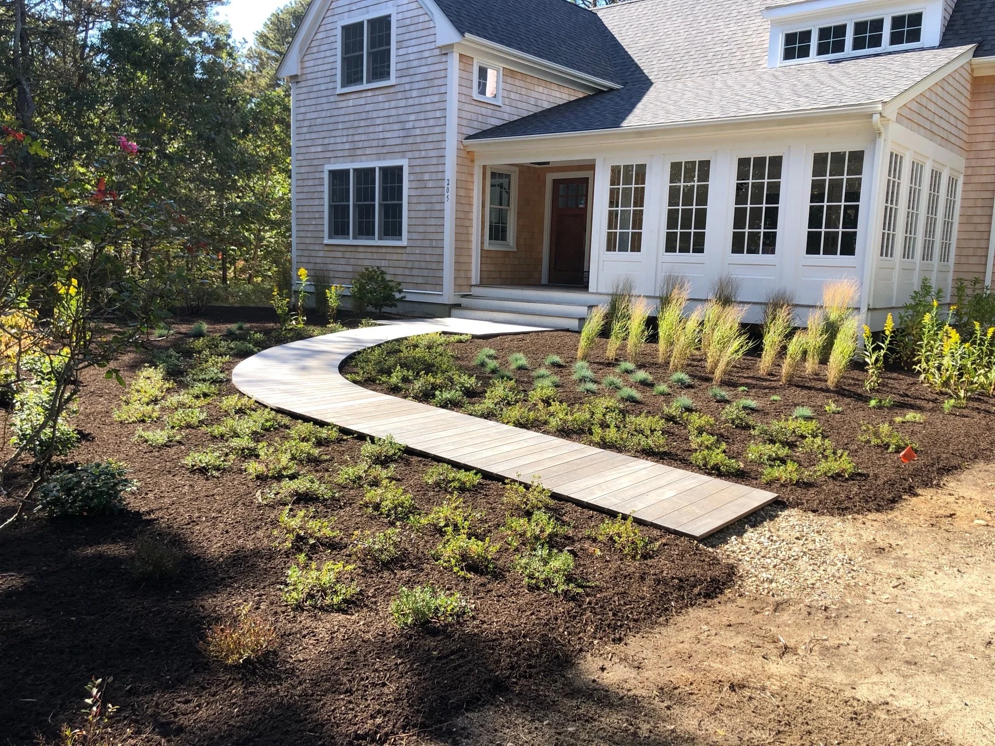 A house with a front porch and landscaped yard featuring a curved wooden walkway, newly planted shrubs, ornamental grasses, and a garden bed with dark mulch.