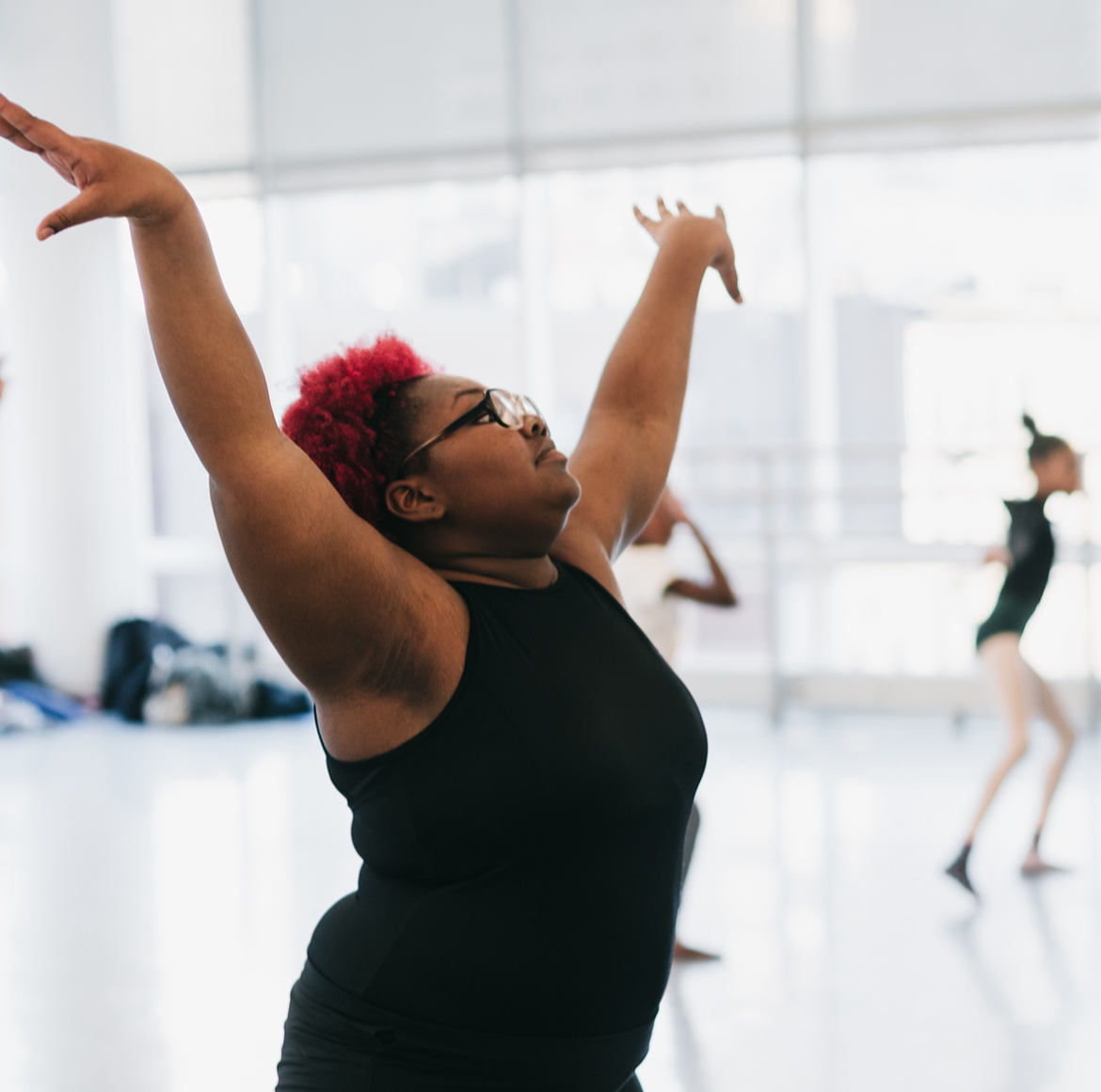 A woman with red hair and glasses stretching with arms raised in a dance or exercise studio with large windows