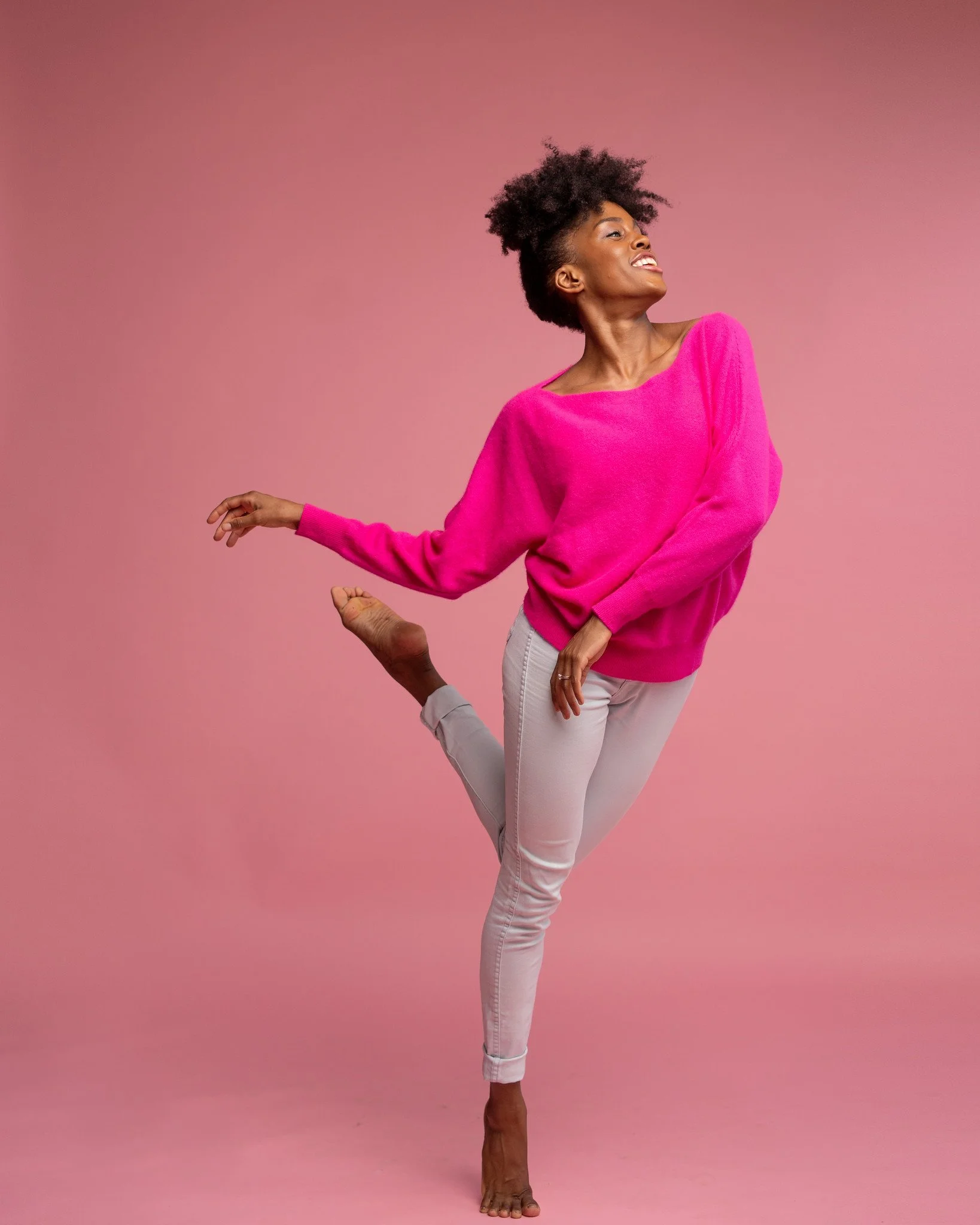 A woman in a bright pink sweater and light-colored pants performing a dance pose against a pink background.