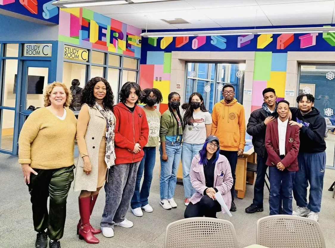 Group of 10 diverse people standing in a colorful room with 'Tigers' rainbow lettering on the wall, some wearing masks, inside a school or community center.
