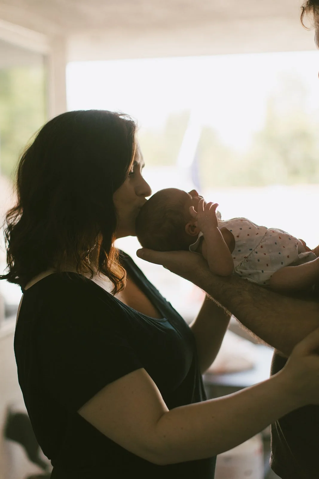 A woman is holding a newborn baby, who is lying on her arm, and kisses the baby's head. The scene is backlit with soft natural light coming from a window in the background.