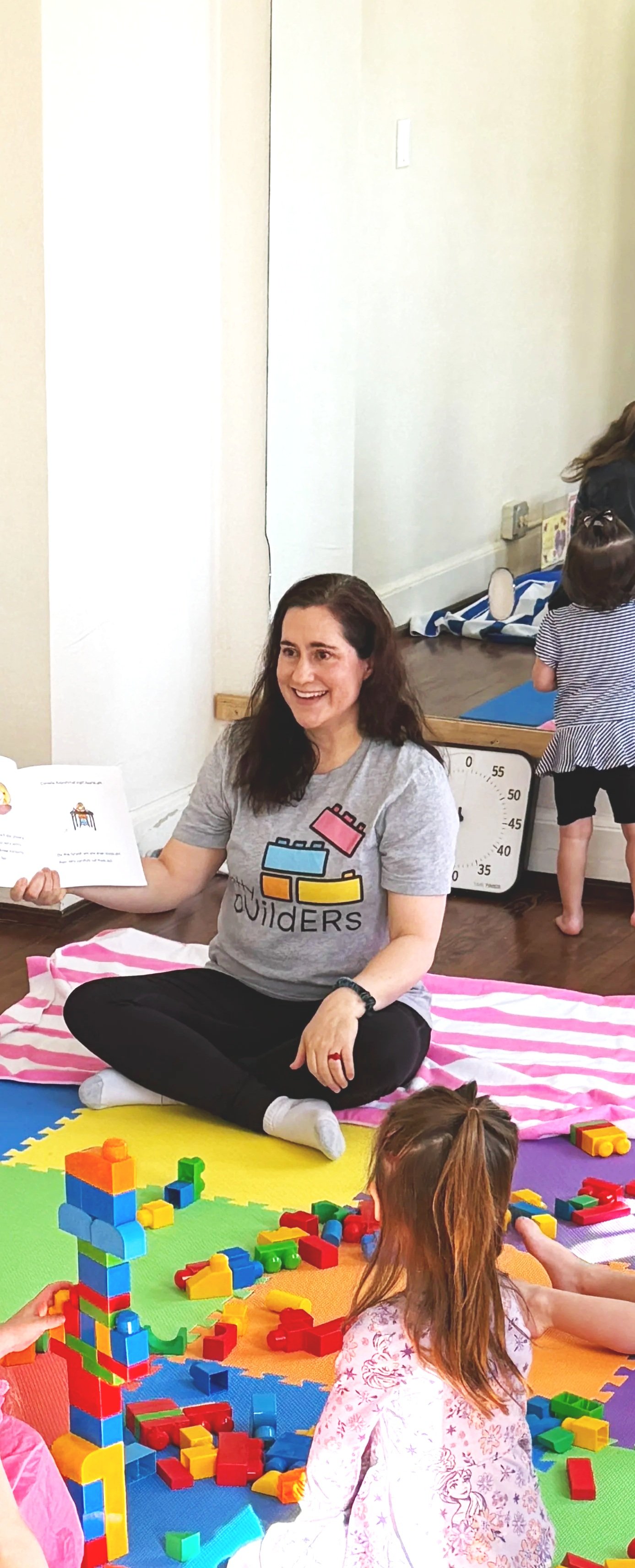 A woman sitting on a colorful play mat in a classroom, reading a book to children. There are toys and building blocks around her, and children are engaged in play.