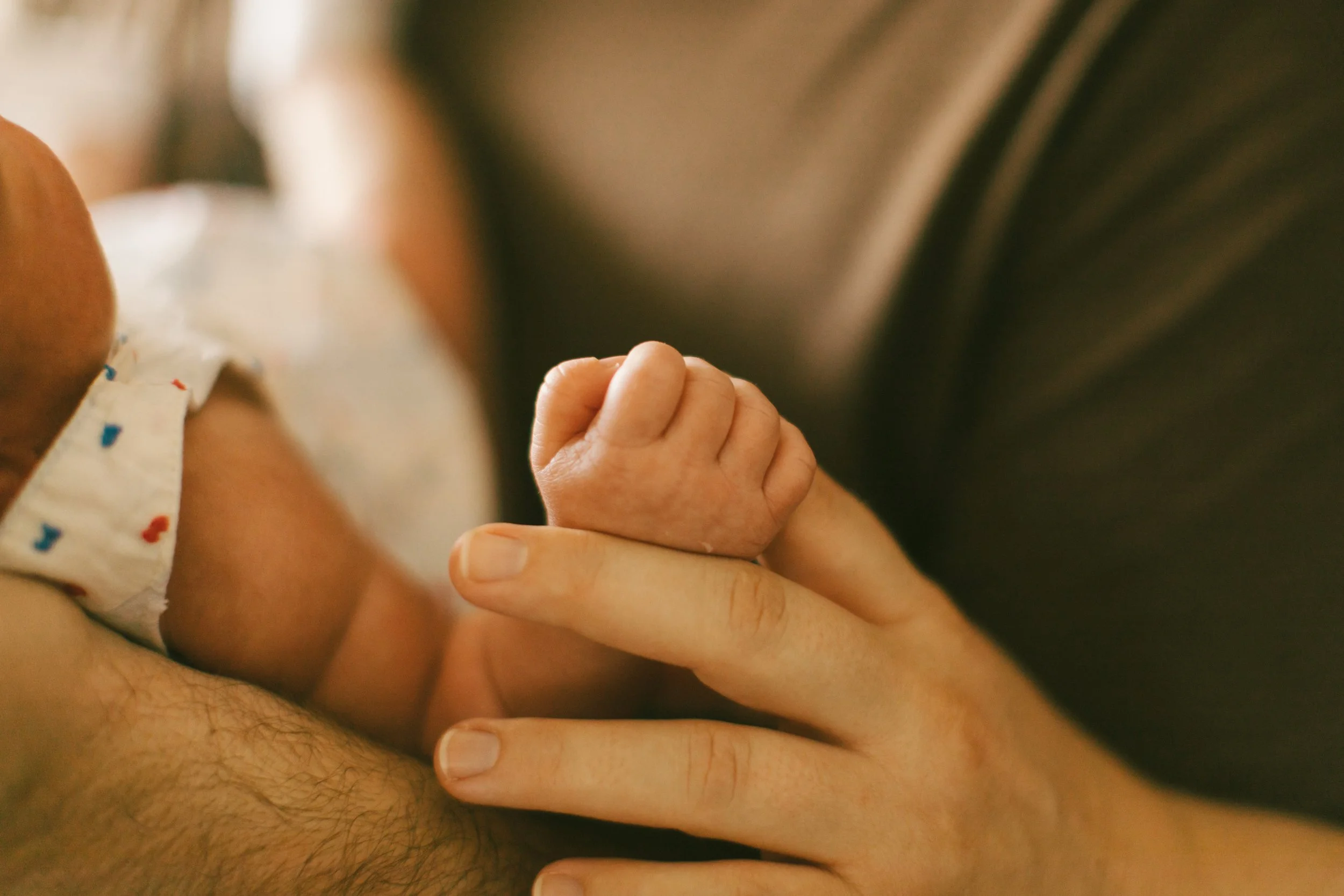 A close-up of a baby’s hand gripping an adult's finger, with the adult’s arm providing support. The background is blurred.