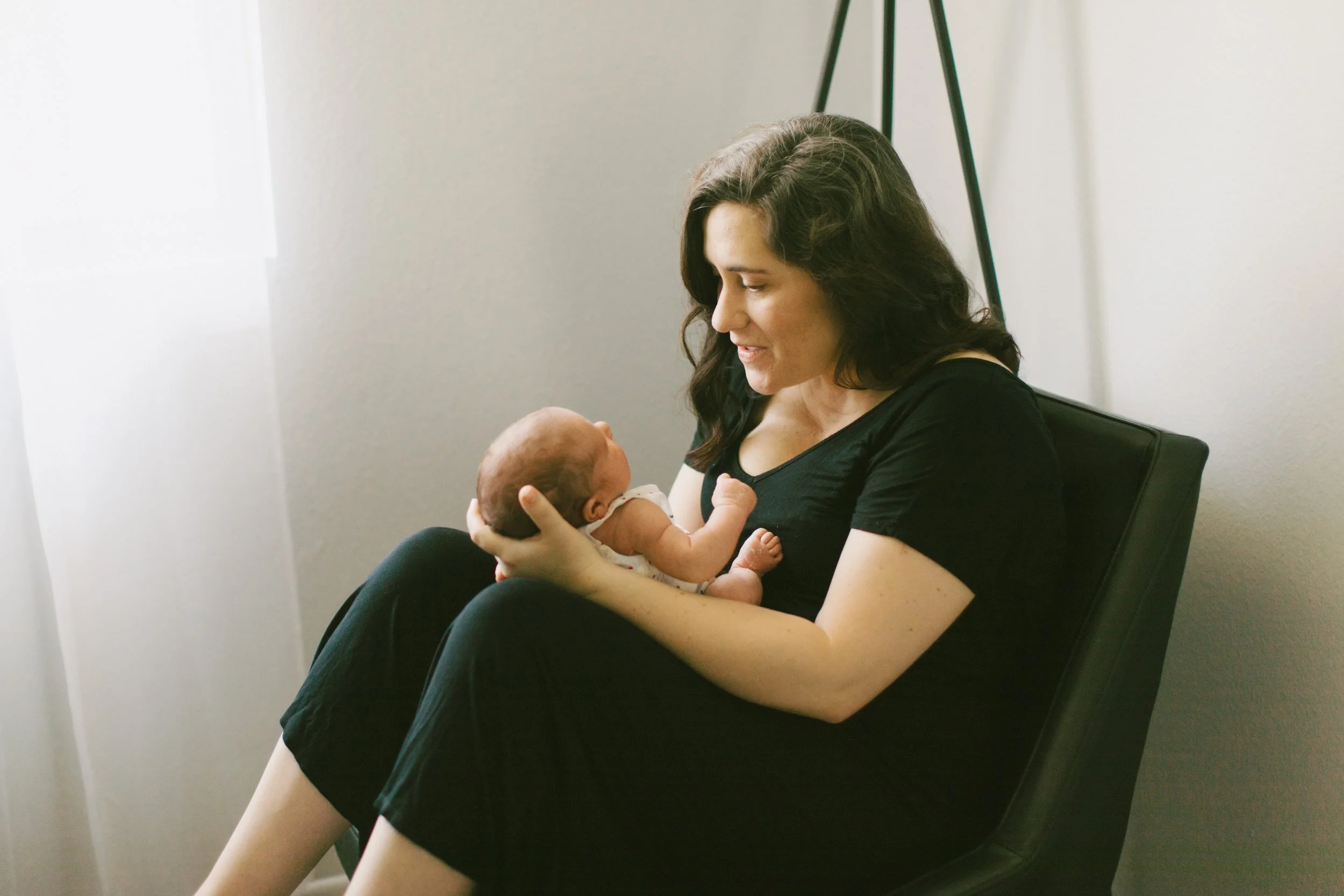 A woman holding a newborn baby in her arms, sitting in a chair with a neutral background.