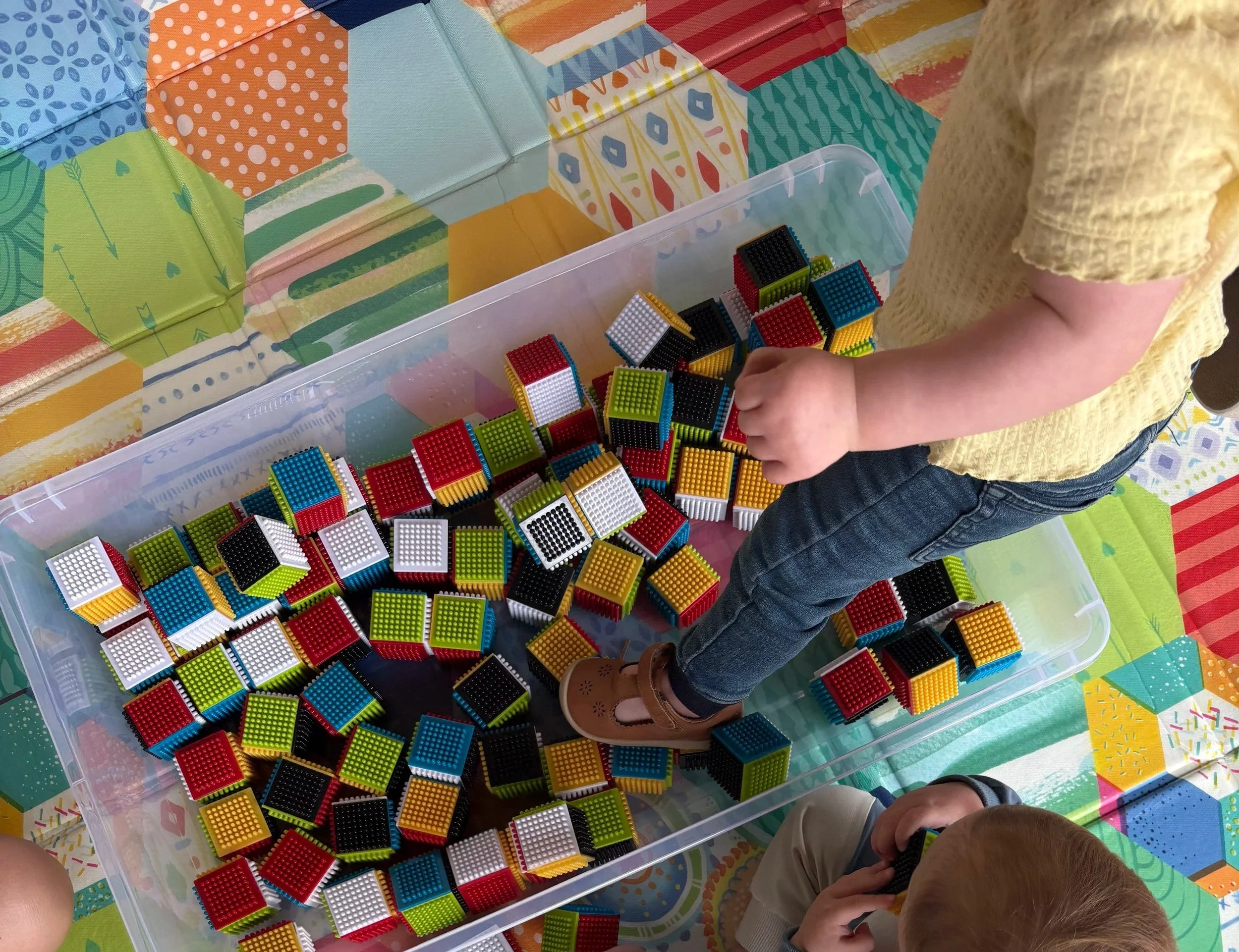 Child playing with multicolored Lego bricks in a plastic container, sitting at a table covered with a colorful, patterned tablecloth.