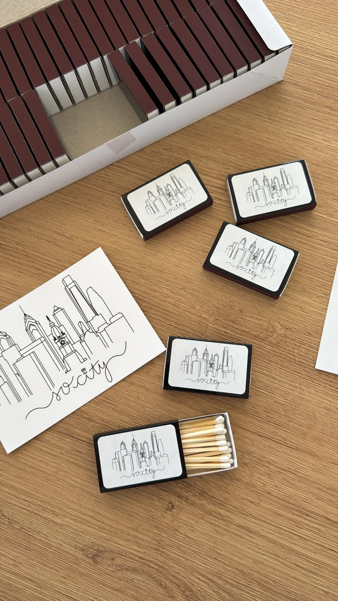 Box of matches with cityscape design and the word "society" on a wooden surface alongside a black-and-white cityscape drawing and other matchboxes.