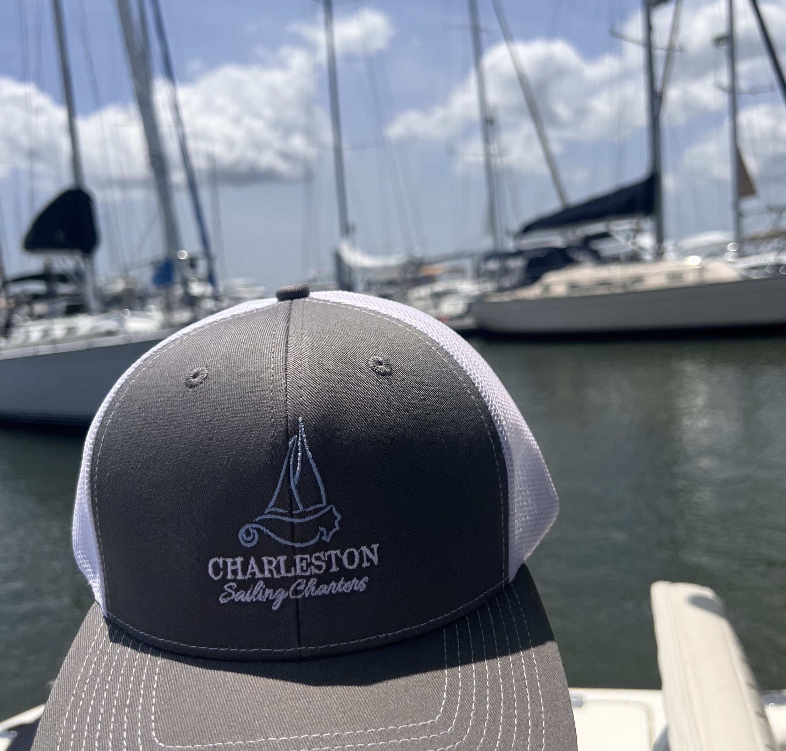 A gray and white Charleston Sailing Charters baseball cap in front of sailboats docked at a marina, with blue sky and clouds in the background.