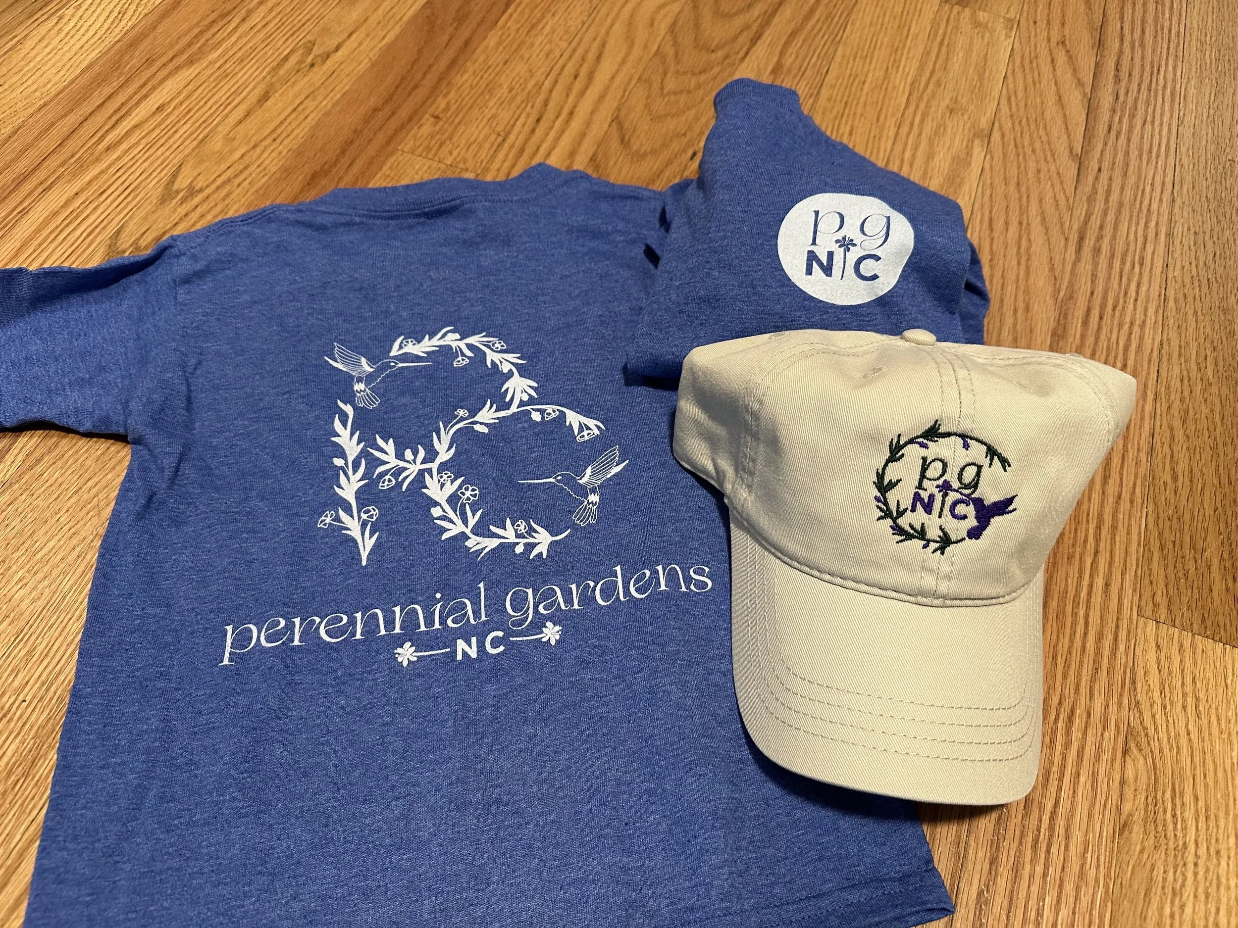 Blue t-shirt with 'perennial gardens NC' logo, beige cap with embroidered 'perennial gardens NC' logo, and a blue baseball cap with 'pg NC' logo on a wooden floor.