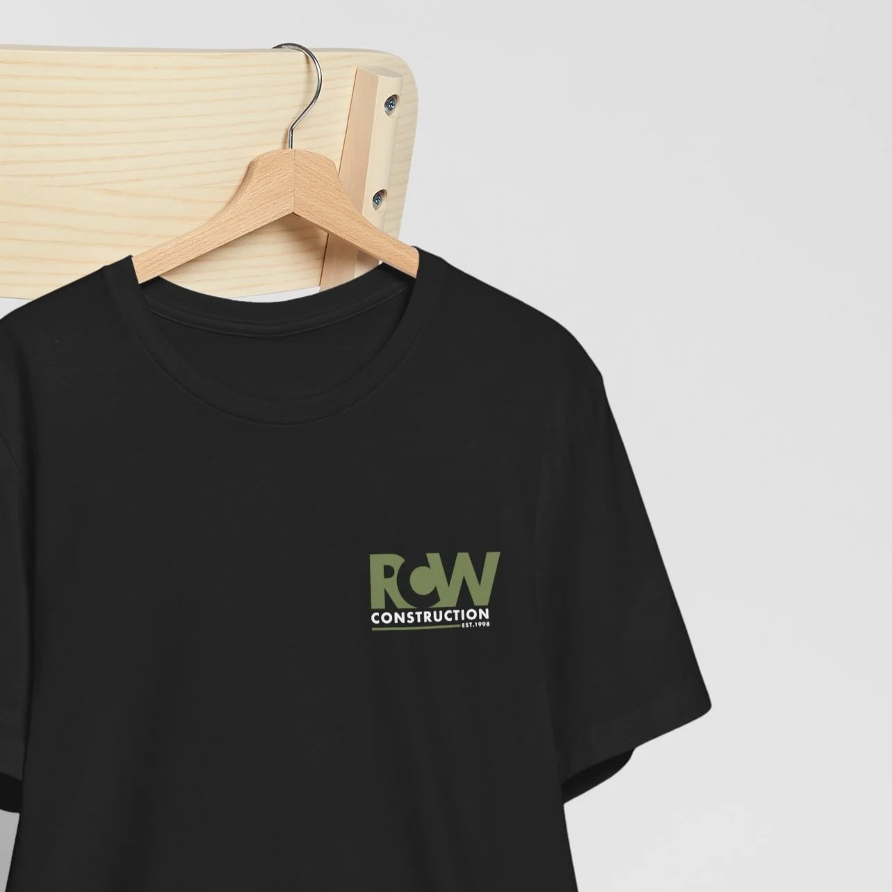 Black T-shirt hanging on a wooden hanger, displaying a logo with the text 'RCW Construction' on it, against a light gray background.