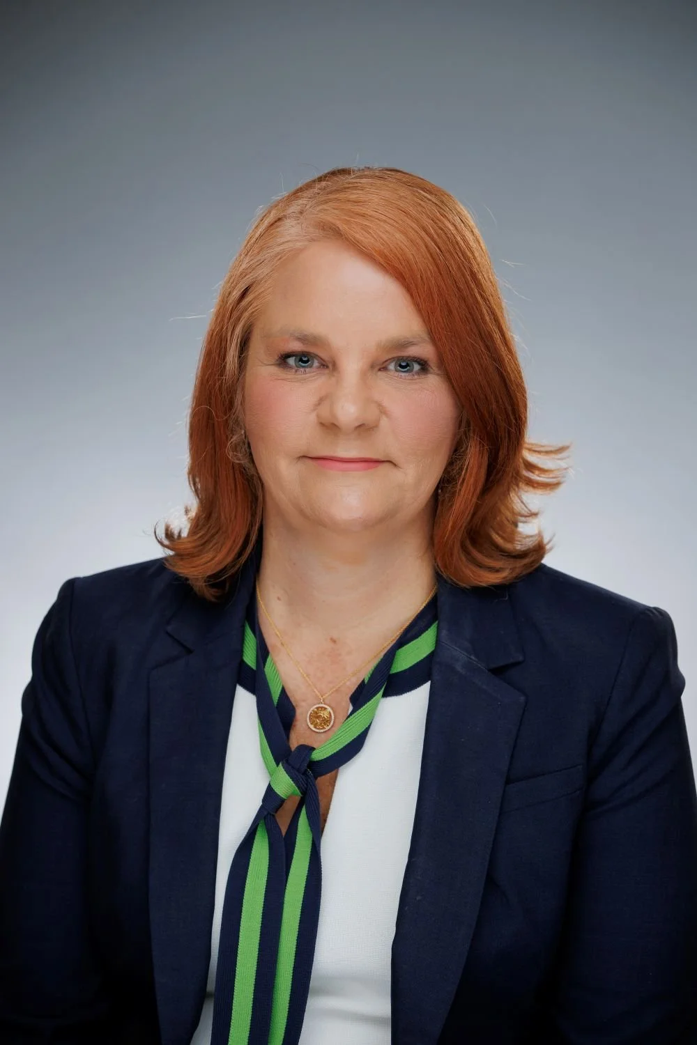 A woman with shoulder-length red hair, wearing a navy blazer, white top, and a blue and green striped scarf, against a gray gradient background.