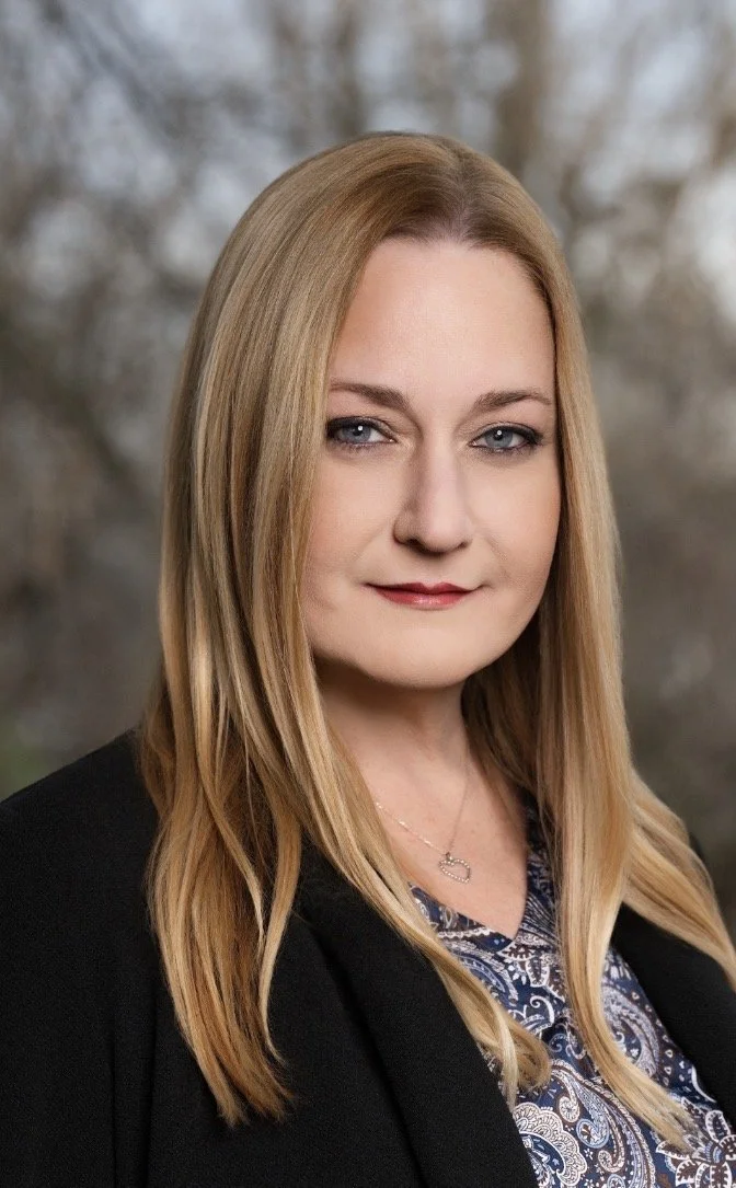 Portrait of a woman with long blonde hair wearing a black blazer and patterned blouse, standing outdoors with blurred trees in the background.
