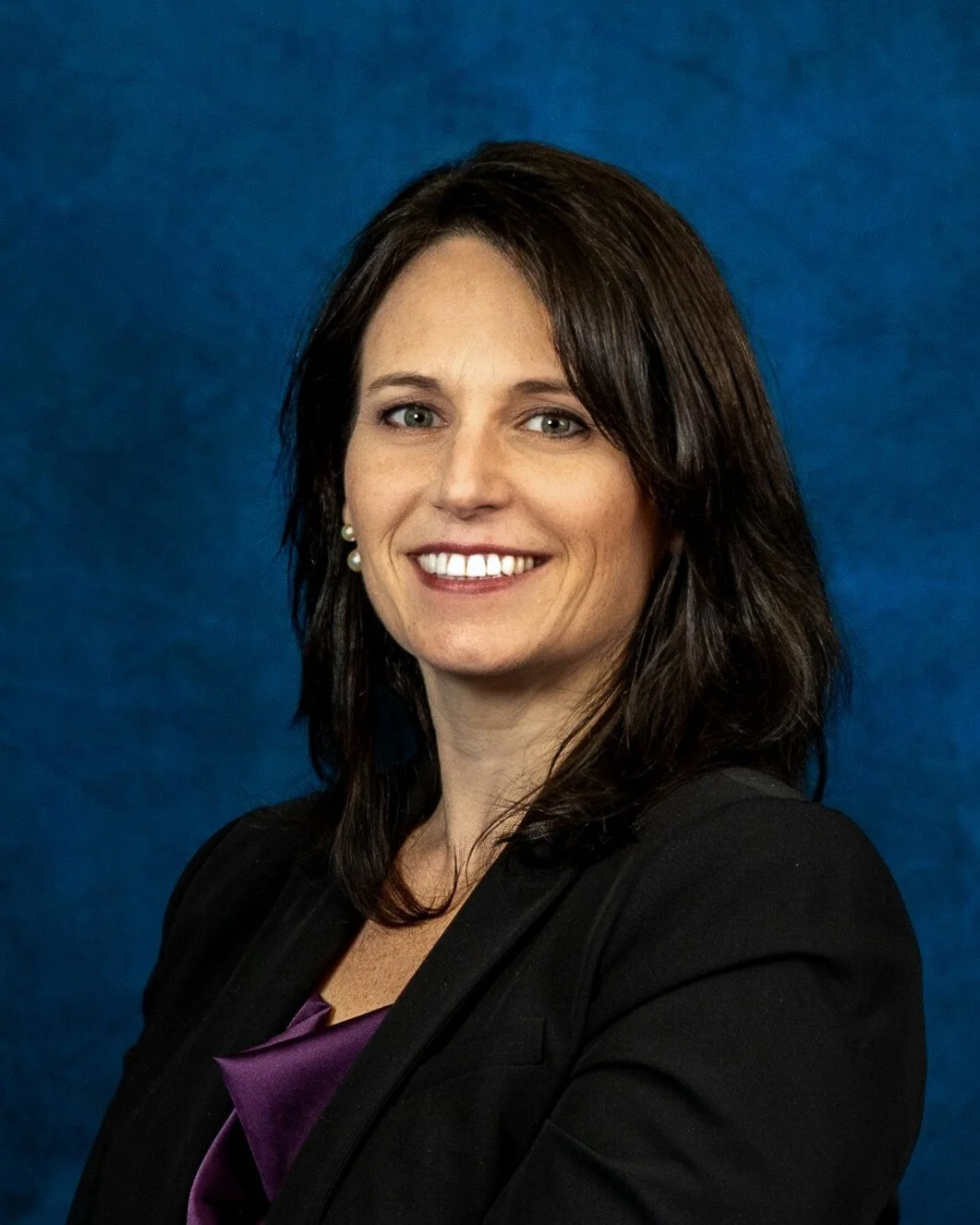 A professional woman with shoulder-length dark brown hair, wearing a black blazer, purple top, pearl earrings, smiling against a blue background.