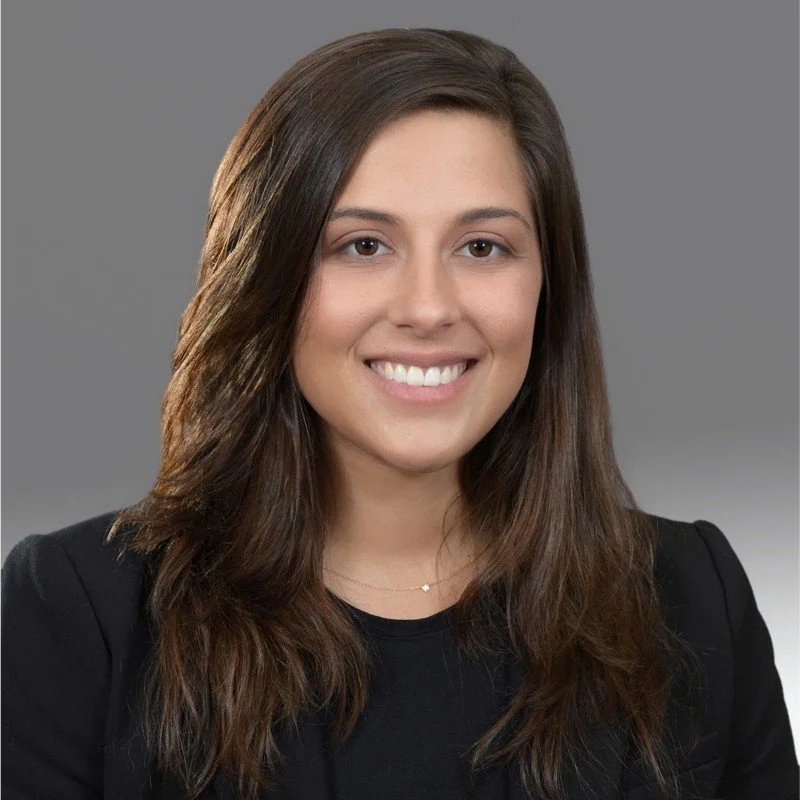 Headshot of a young woman with long, dark brown hair, smiling, wearing a black blazer and a delicate necklace, against a neutral gray background.
