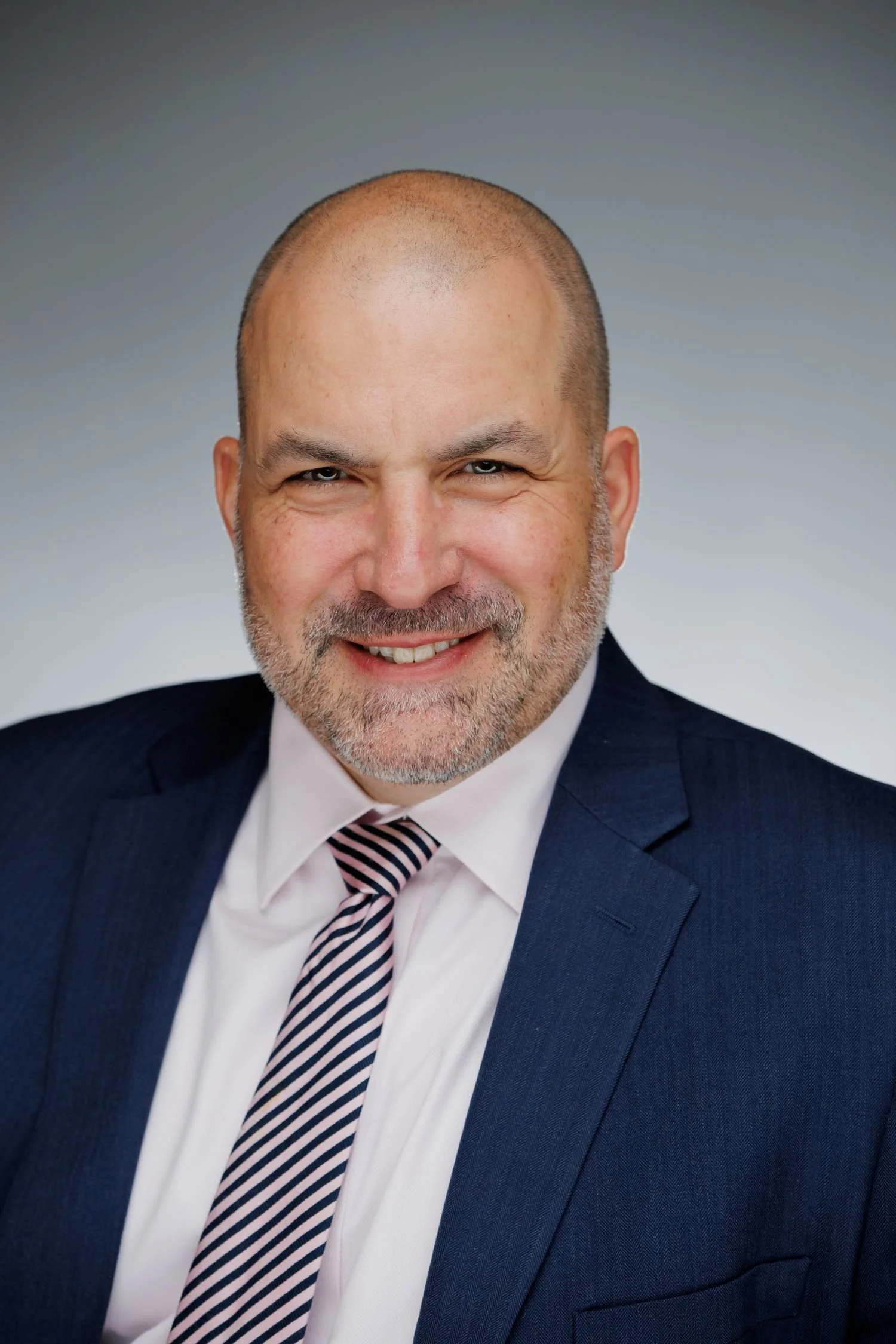 Professional headshot of a smiling man with a beard, wearing a dark suit, white shirt, and a striped tie.
