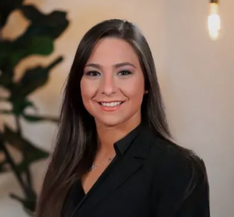 A young woman with long dark hair, smiling, wearing a black top, in an indoor setting with a plant in the background.