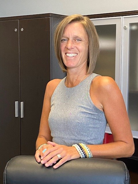 Beth Chambers with shoulder-length light brown hair wearing a sleeveless gray top, smiling and standing behind a black chair in an office or conference room.
