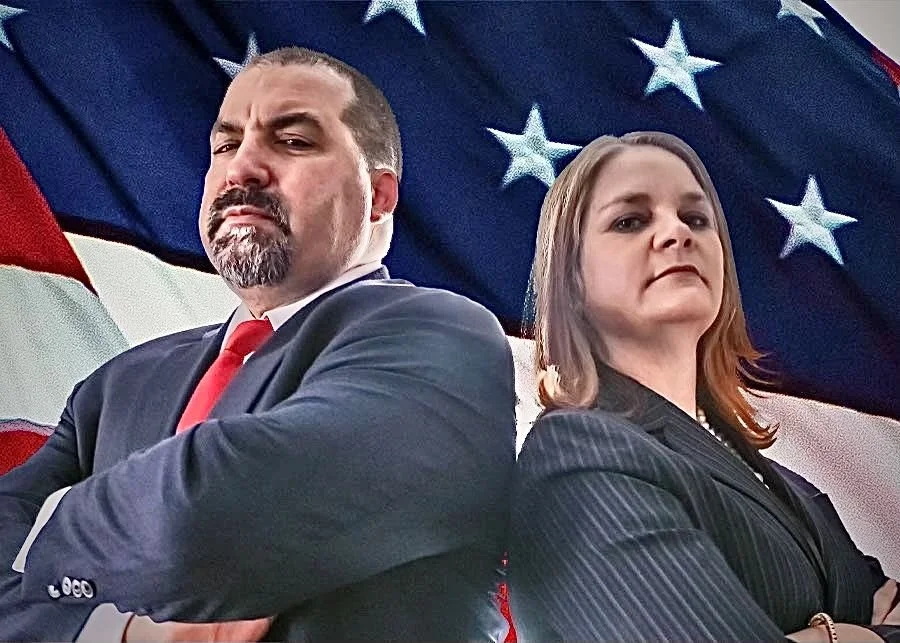 A man and woman stand back to back in front of an American flag, with serious expressions and arms crossed.