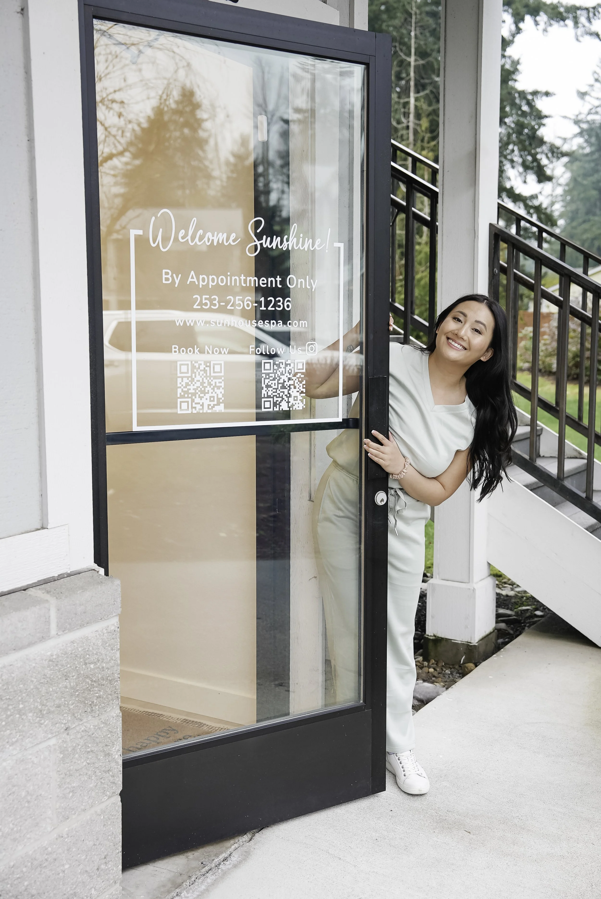 A smiling woman peeking out from behind a glass door with the sign, 'Welcome Sunshine, By Appointment Only, 253-256-1236, www.sunhousespab.com, Book Now, Follow Us' on it.