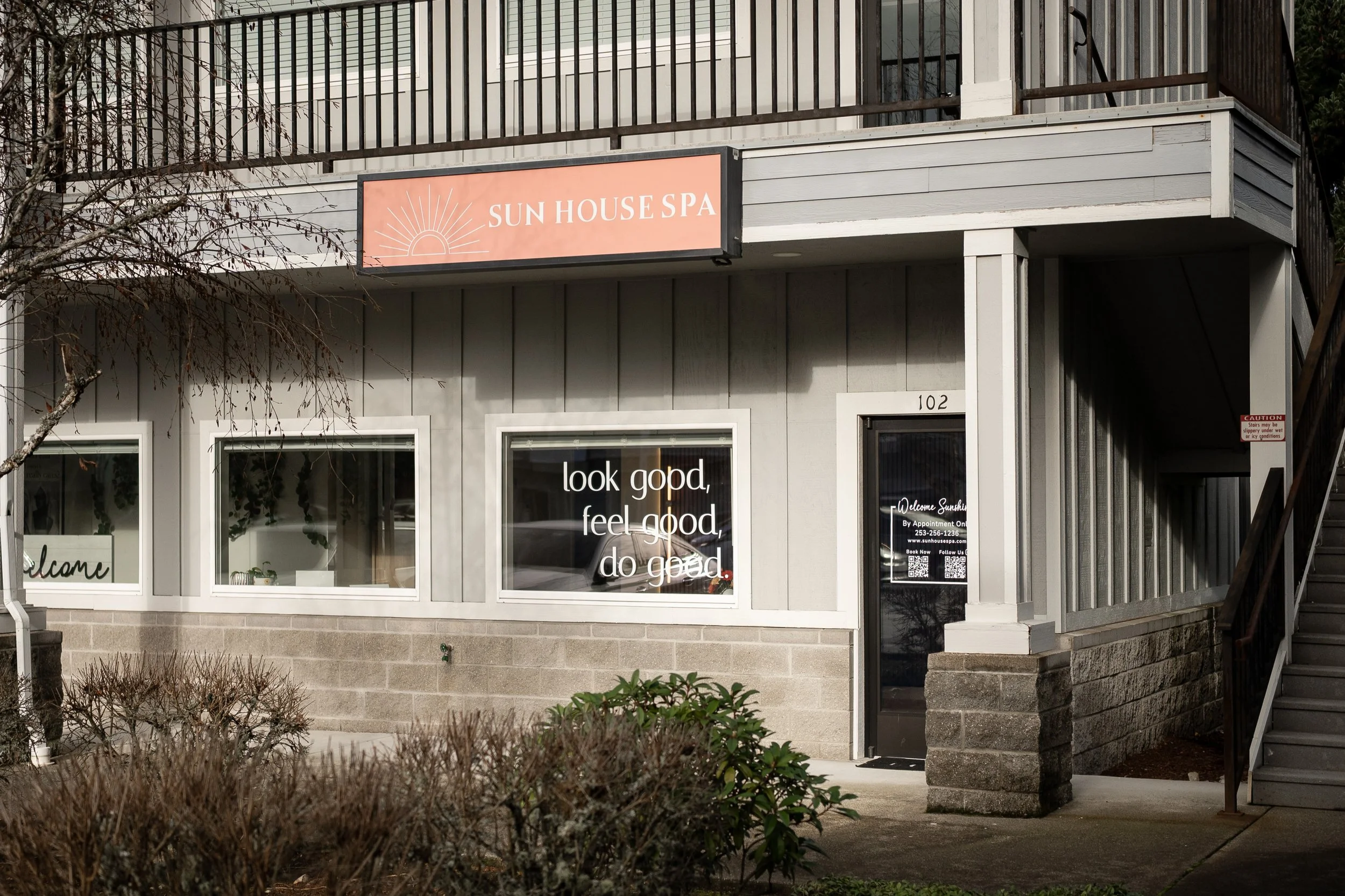 Exterior of a spa named 'Sun House Spa' with a sign, windows, and a door, surrounded by bushes and a staircase.