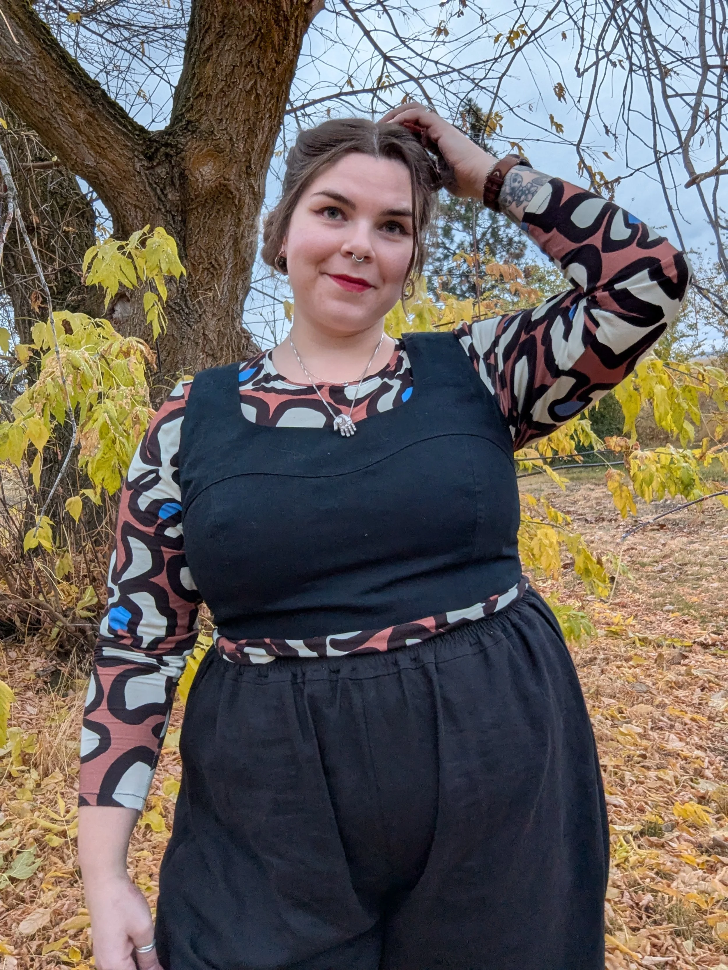 A whited woman with light brown hair and red lipstick smiles with one hand in her hair. She is wearing black pants, a brown, blue, white and black floral printed long sleeve top with a black corset style tank over top.