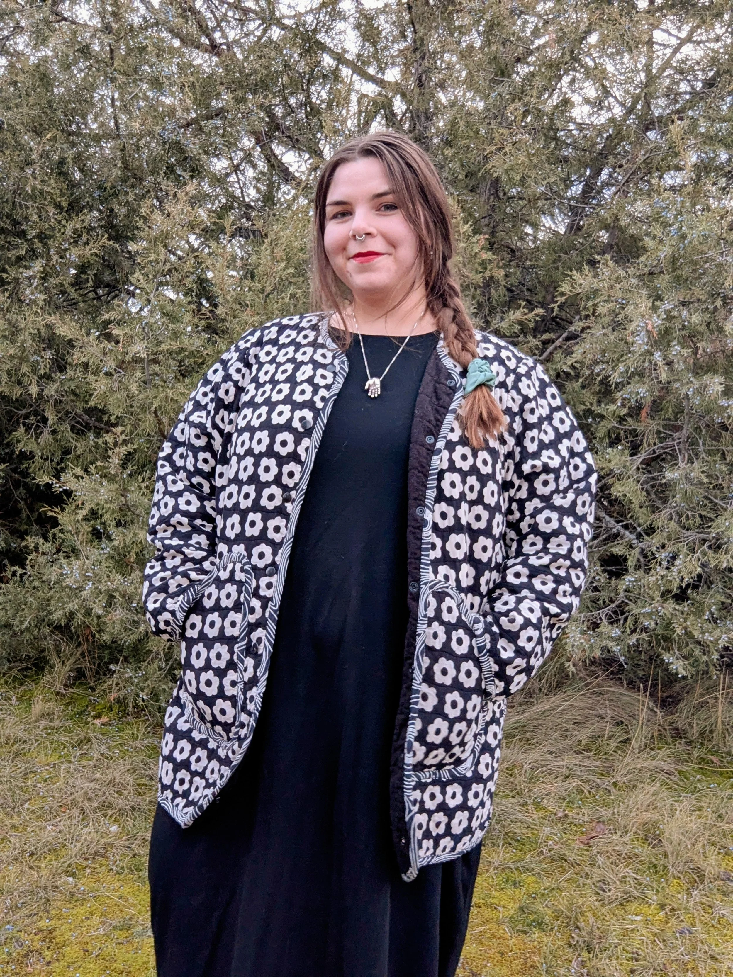 A white woman with light brown hair and red lipstick smiles gently. She is wearing silver jewelry, black dress and a black and white floral hand made quilt coat.