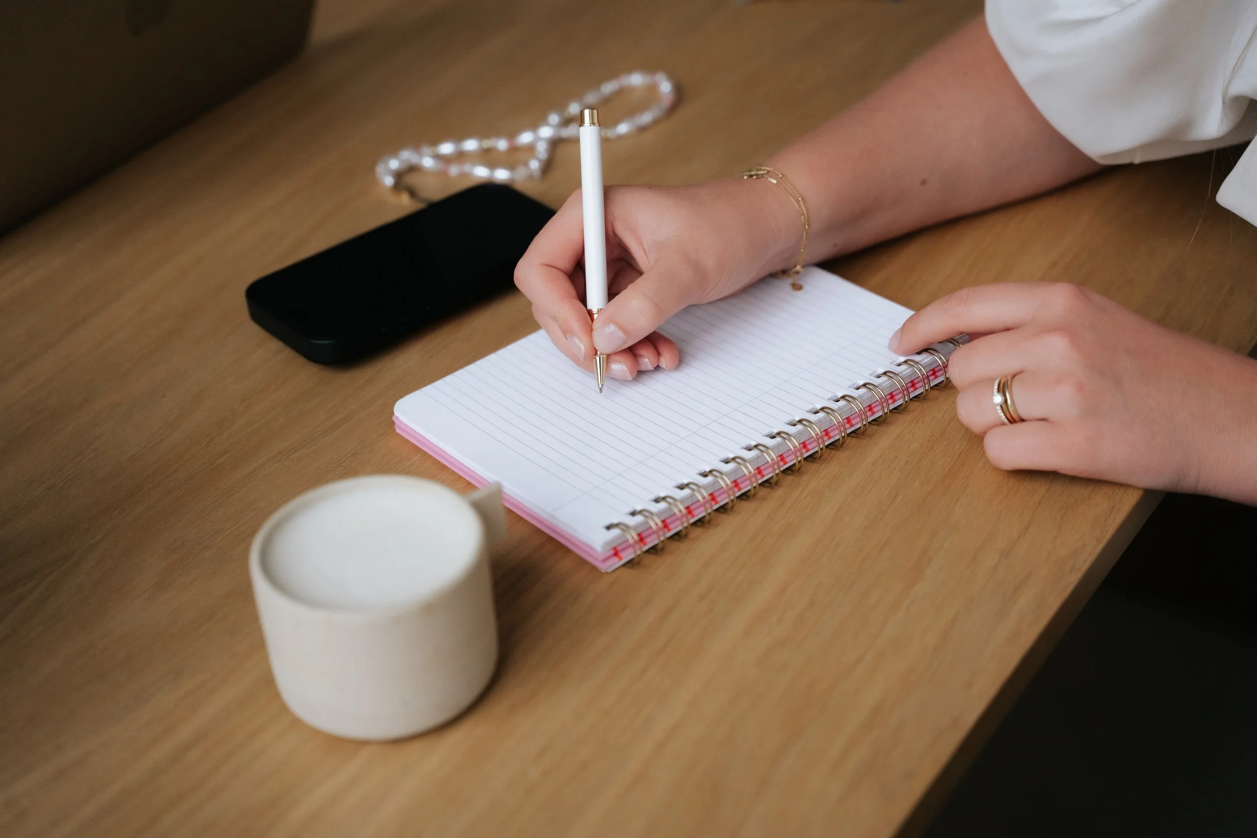 Person writing in a spiral notebook with a white pen on a wooden table. There is a black phone, a white mug, and a beaded necklace nearby.