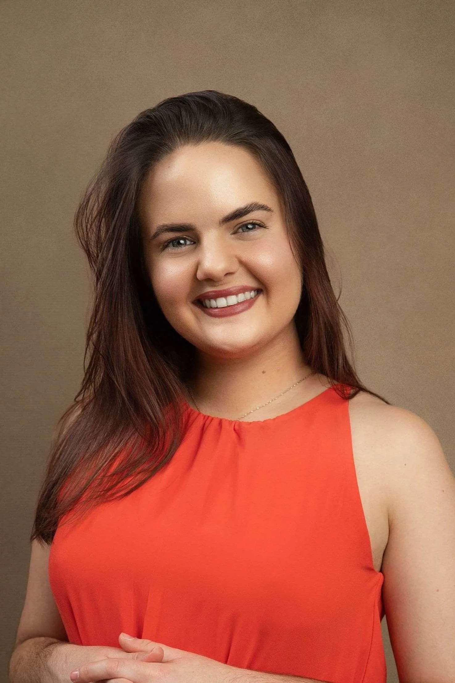 Ericka Vargas Headshot: young Hispanic female therapist with long brown hair smiling outdoors, wearing an orange sleeveless top, with green foliage in the background.