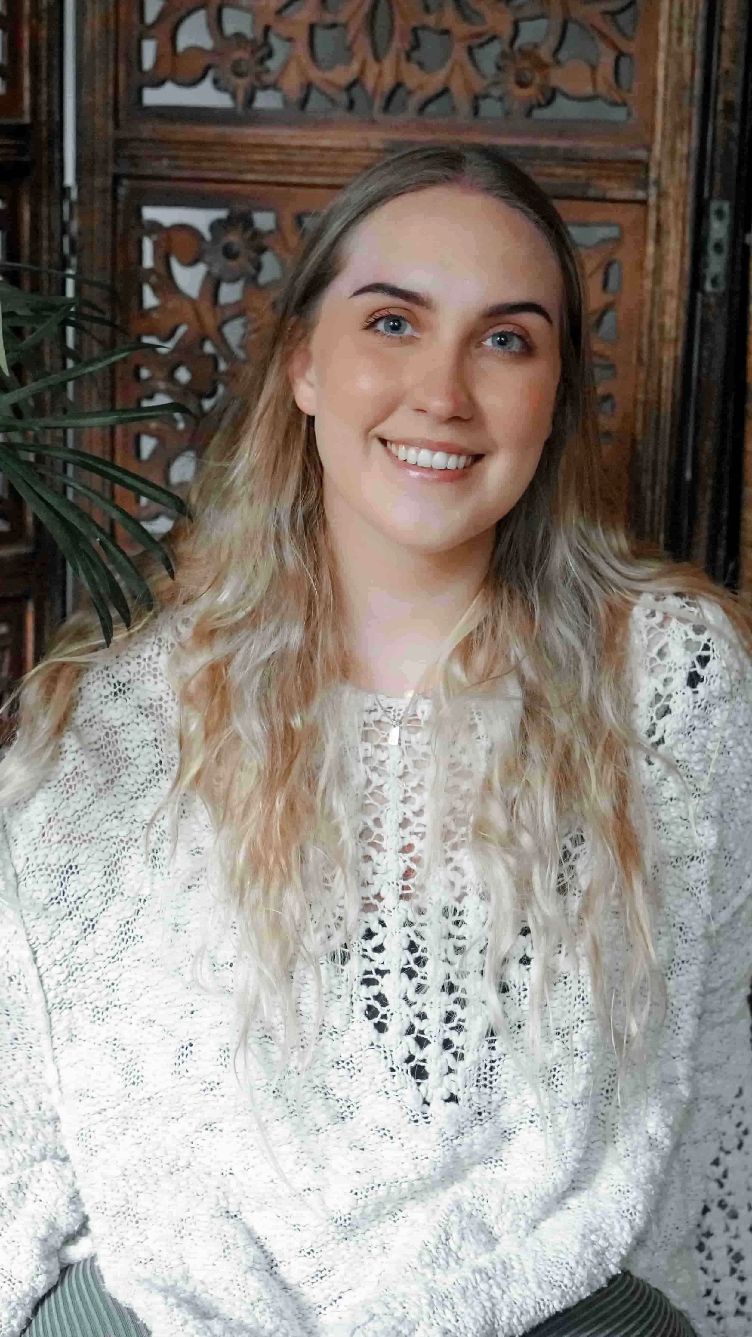 Taci Clay Headshot: A young female therapist with long, wavy blonde hair and blue eyes smiling at the camera, wearing a white lace blouse and is sitting in front of a wooden decorative background with a leafy green plant partially visible on her left