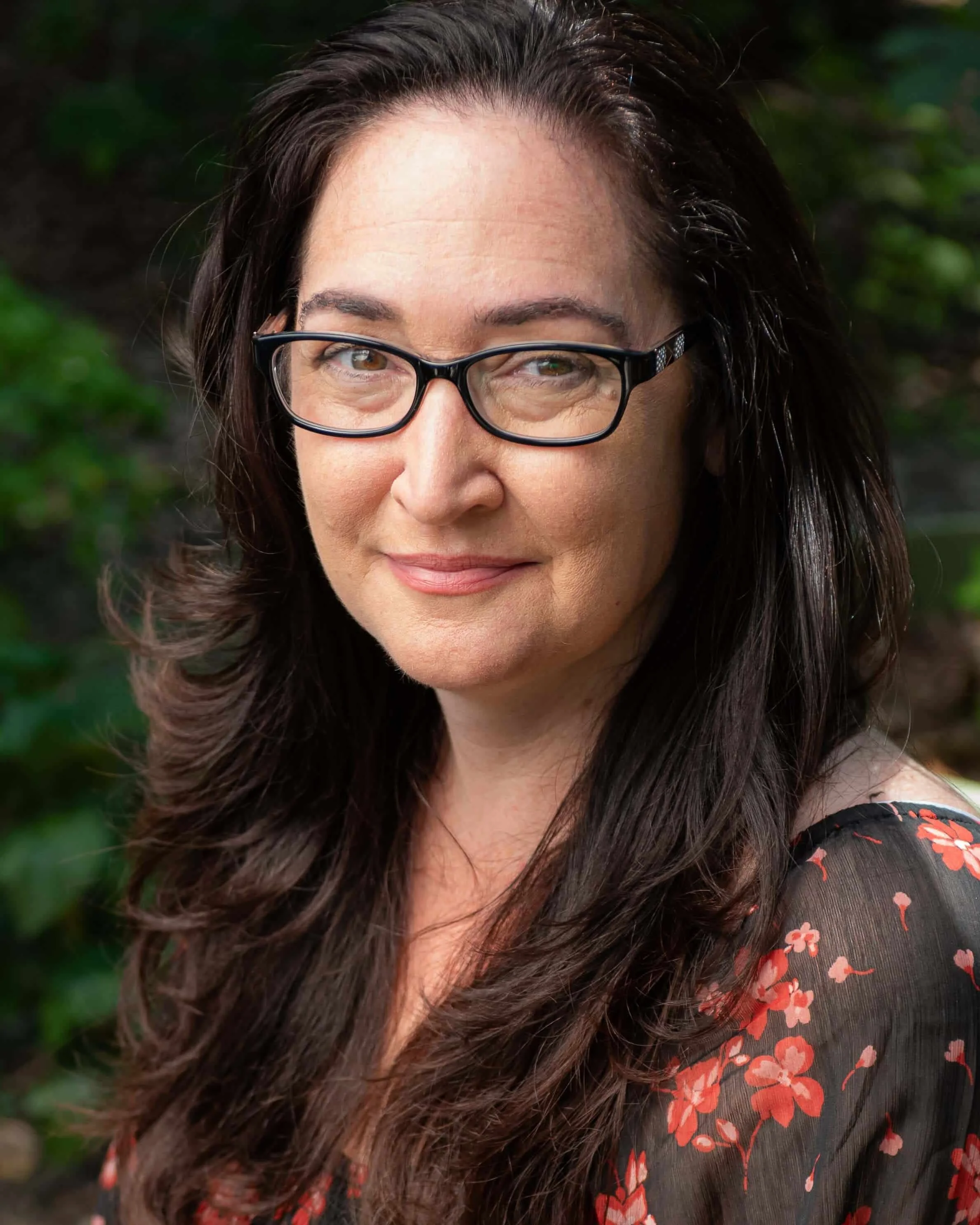 Tracy Bohannon Headshot: A female therapist with long dark hair, glasses, and a slight smile, wearing a black floral top, outdoors with greenery in the background.