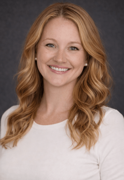 Erika Lowery Headshot: Female therapist in long sleave, long strawberry blond hair, white shirt open mouth smiles at camera, solid gray background