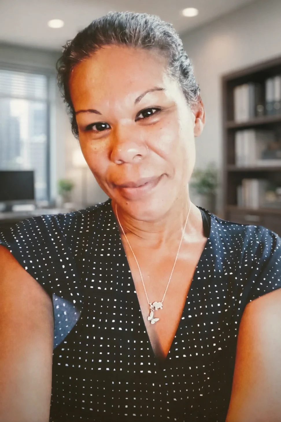 Kim Walker Moore Headshot: Black Female therapist smiling softly at camera, wearing a black shirt with white poka dots, blurred background of an office space
