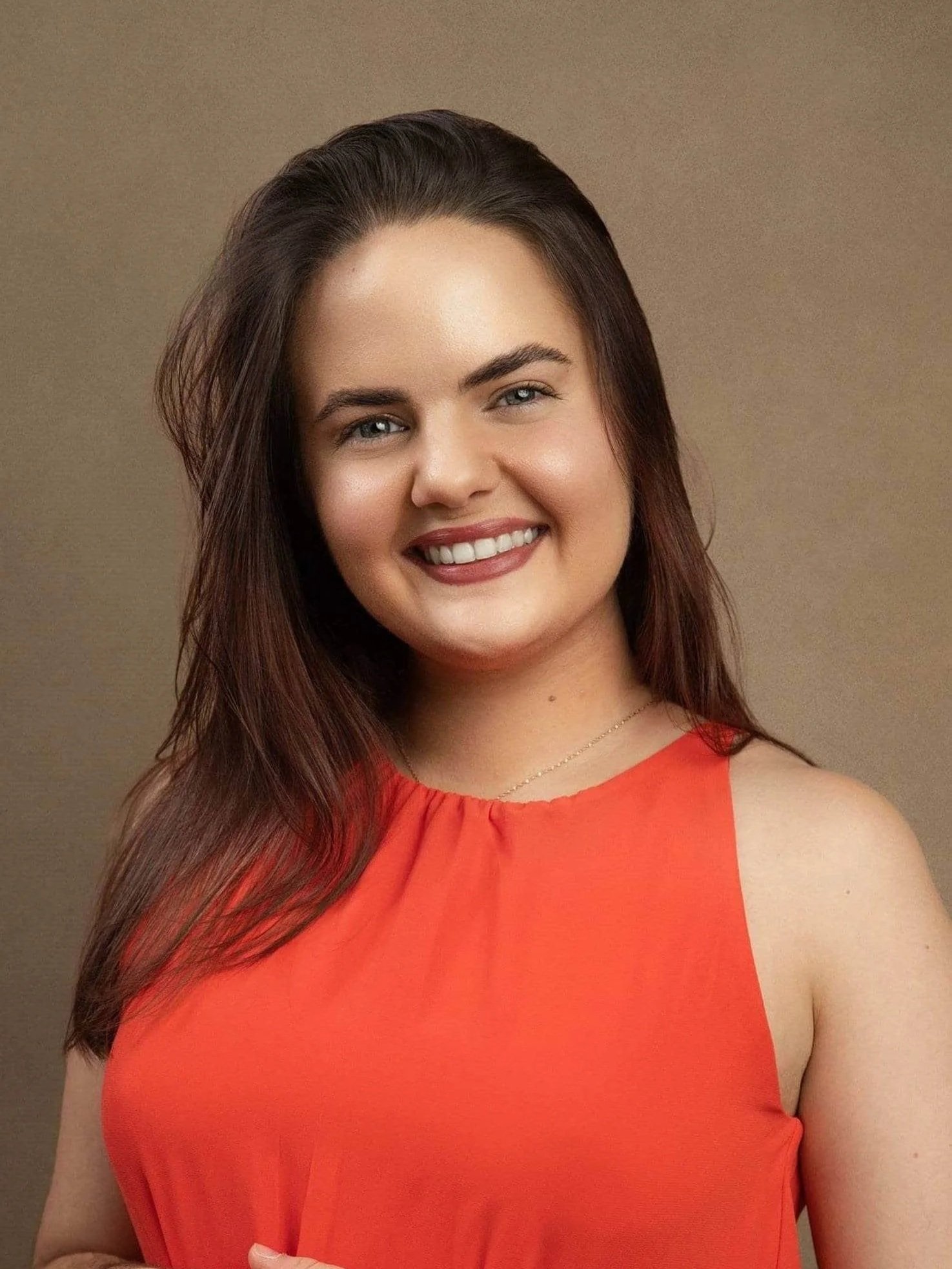 Ericka Vargas Headshot: young Hispanic female therapist with long brown hair smiling outdoors, wearing an orange sleeveless top, with green foliage in the background.
