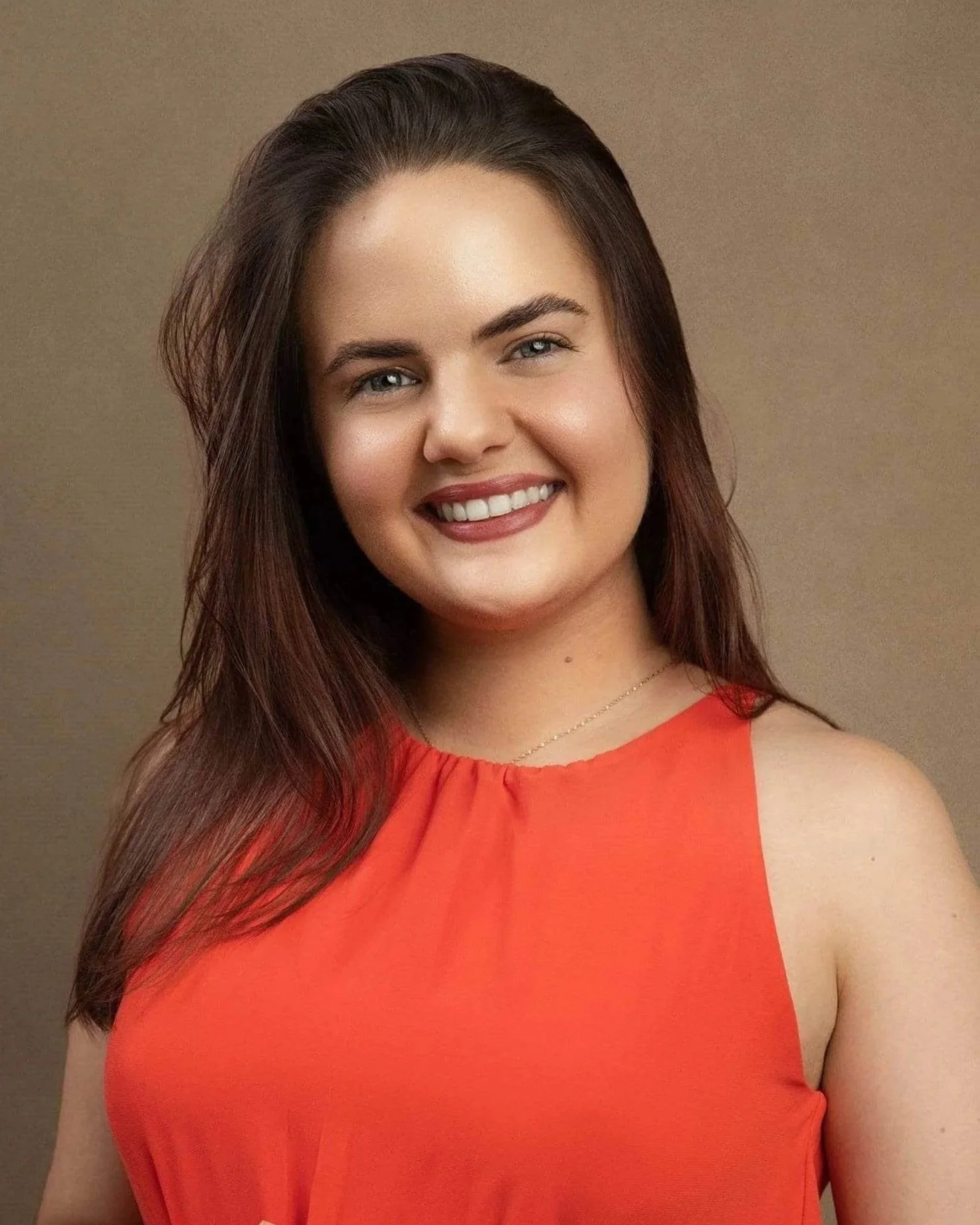Ericka Vargas Headshot: young Hispanic female therapist with long brown hair smiling outdoors, wearing an orange sleeveless top, with green foliage in the background.