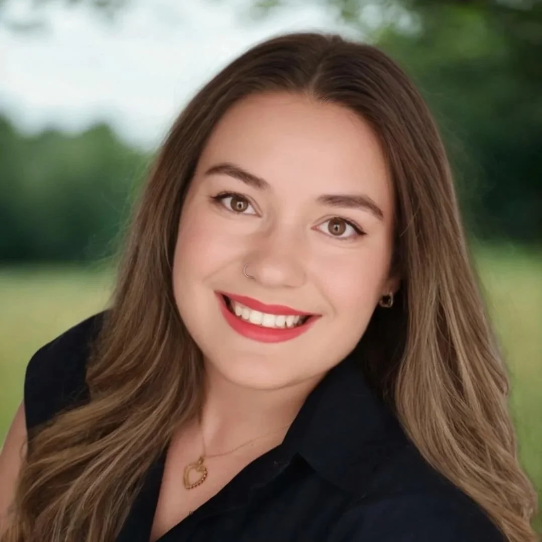 Kristen Johns Headshot: smiling young female therapist with long wavy brown hair, wearing a black top, gold necklace, and earrings, outdoors with blurred greenery in the background.