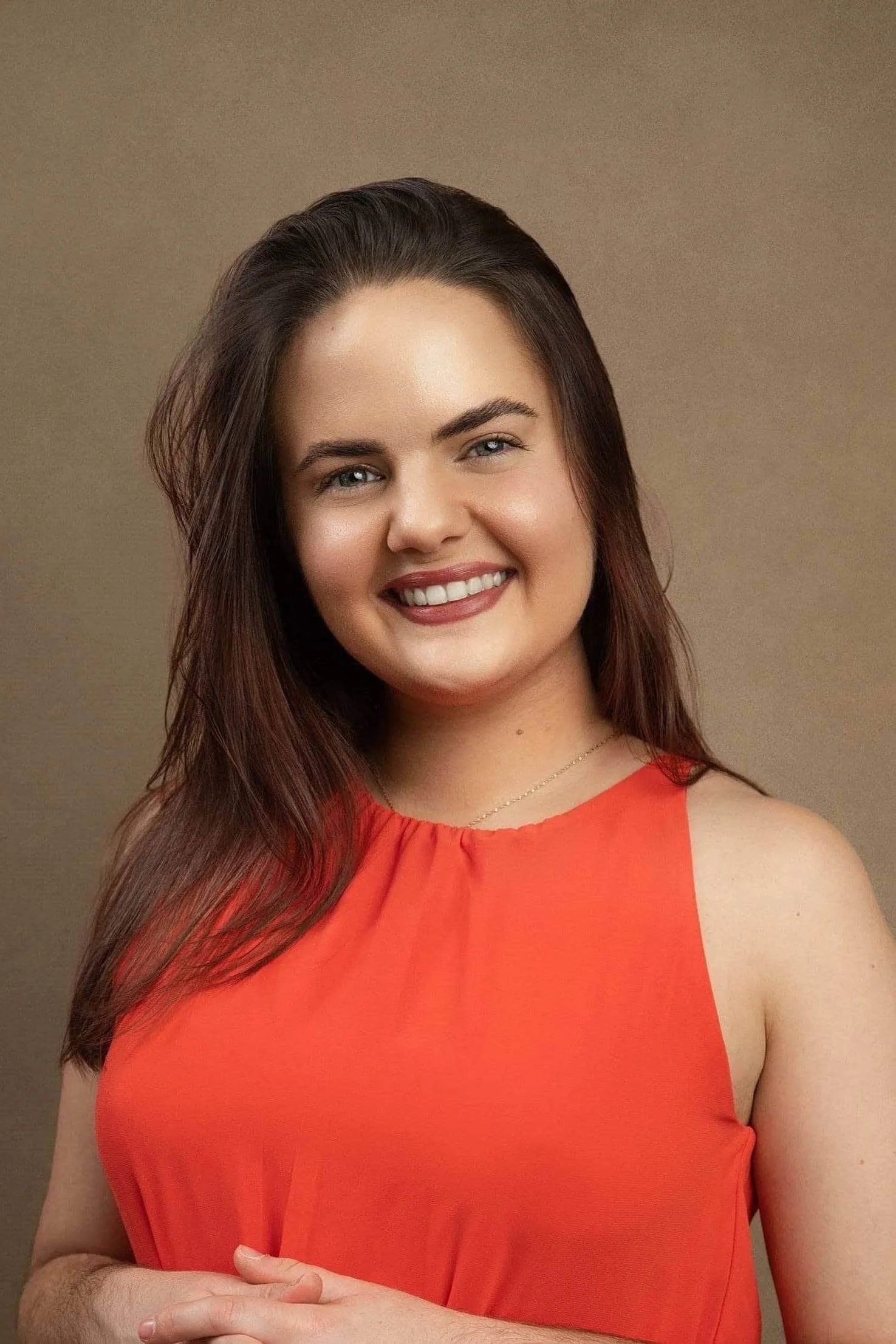 Ericka Vargas Headshot: young Hispanic female therapist with long brown hair smiling outdoors, wearing an orange sleeveless top, with green foliage in the background.