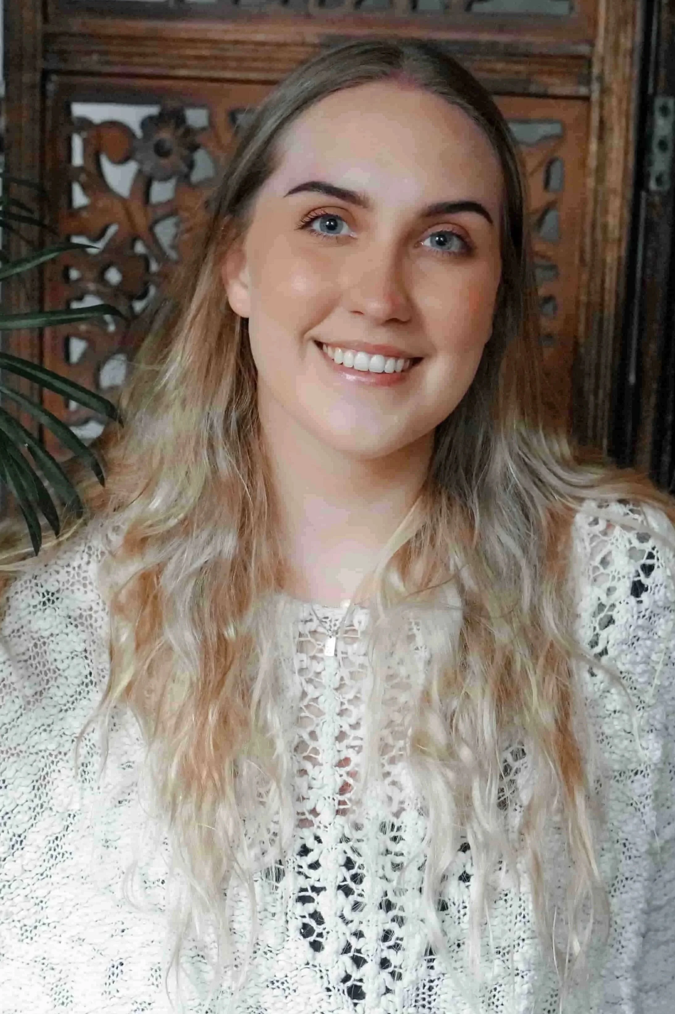 Taci Clay Headshot: A young female therapist with long, wavy blonde hair and blue eyes smiling at the camera, wearing a white lace blouse and is sitting in front of a wooden decorative background with a leafy green plant partially visible on her left