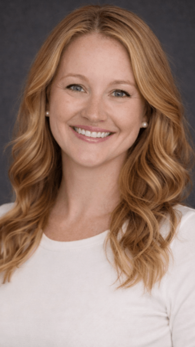 Erika Lowery Headshot: Female therapist in long sleave, long strawberry blond hair, white shirt open mouth smiles at camera, solid gray background