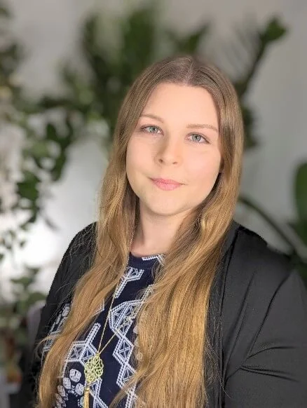 Lauren Cunningwood Headshot: A female therapist with long, light brown hair and blue eyes, smiling gently, wearing a black blazer and a patterned top with a gold necklace, sitting in front of green leafy plants.