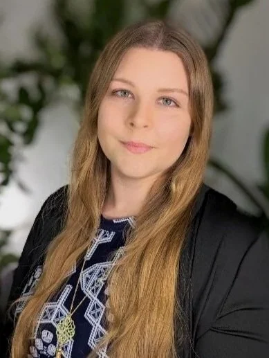 Lauren Cunningwood Headshot: A female therapist with long, light brown hair and blue eyes, smiling gently, wearing a black blazer and a patterned top with a gold necklace, sitting in front of green leafy plants.