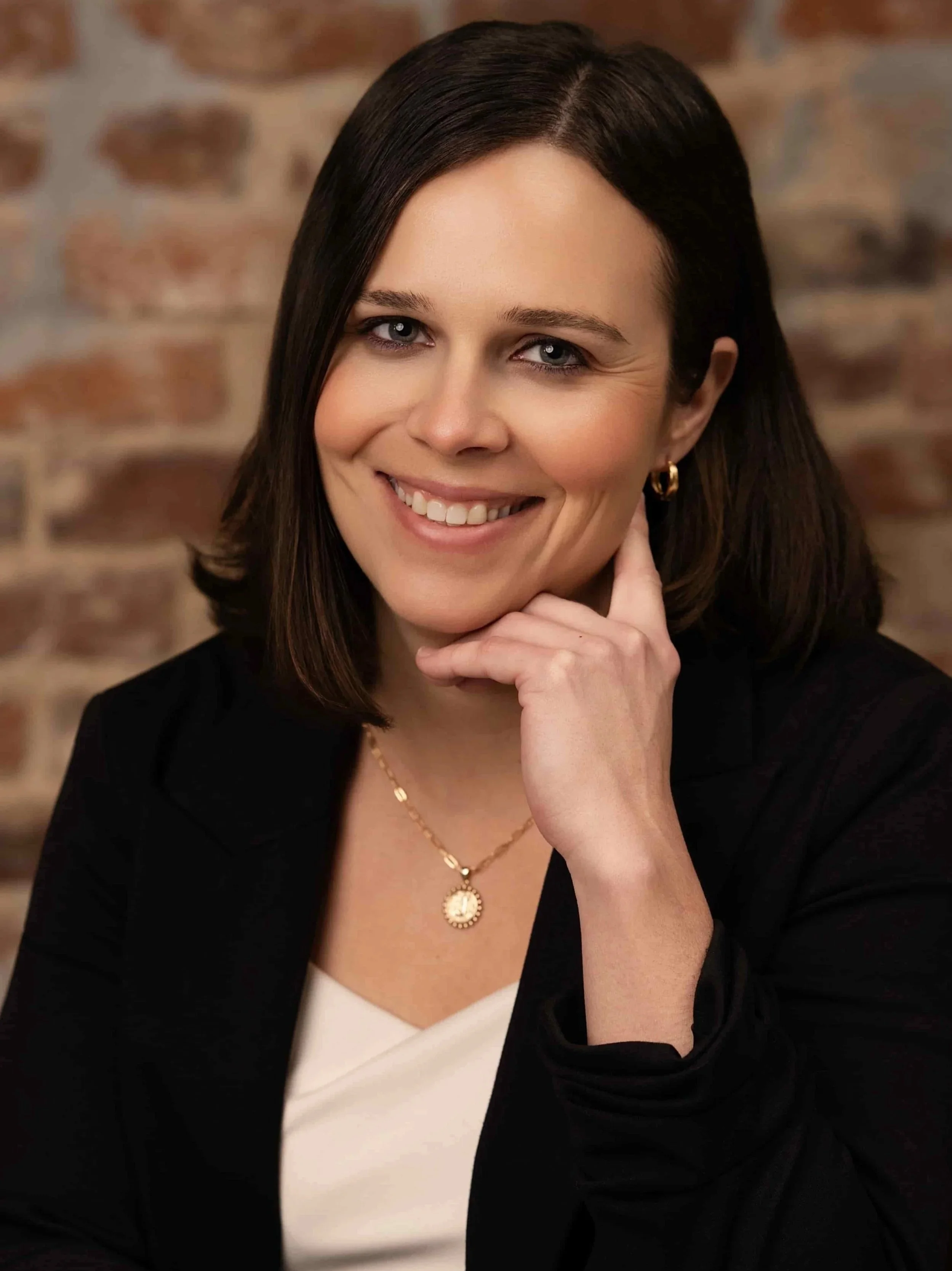 Young female therapist wearing a black blazer and white undershirt smiles at camera with a brick background