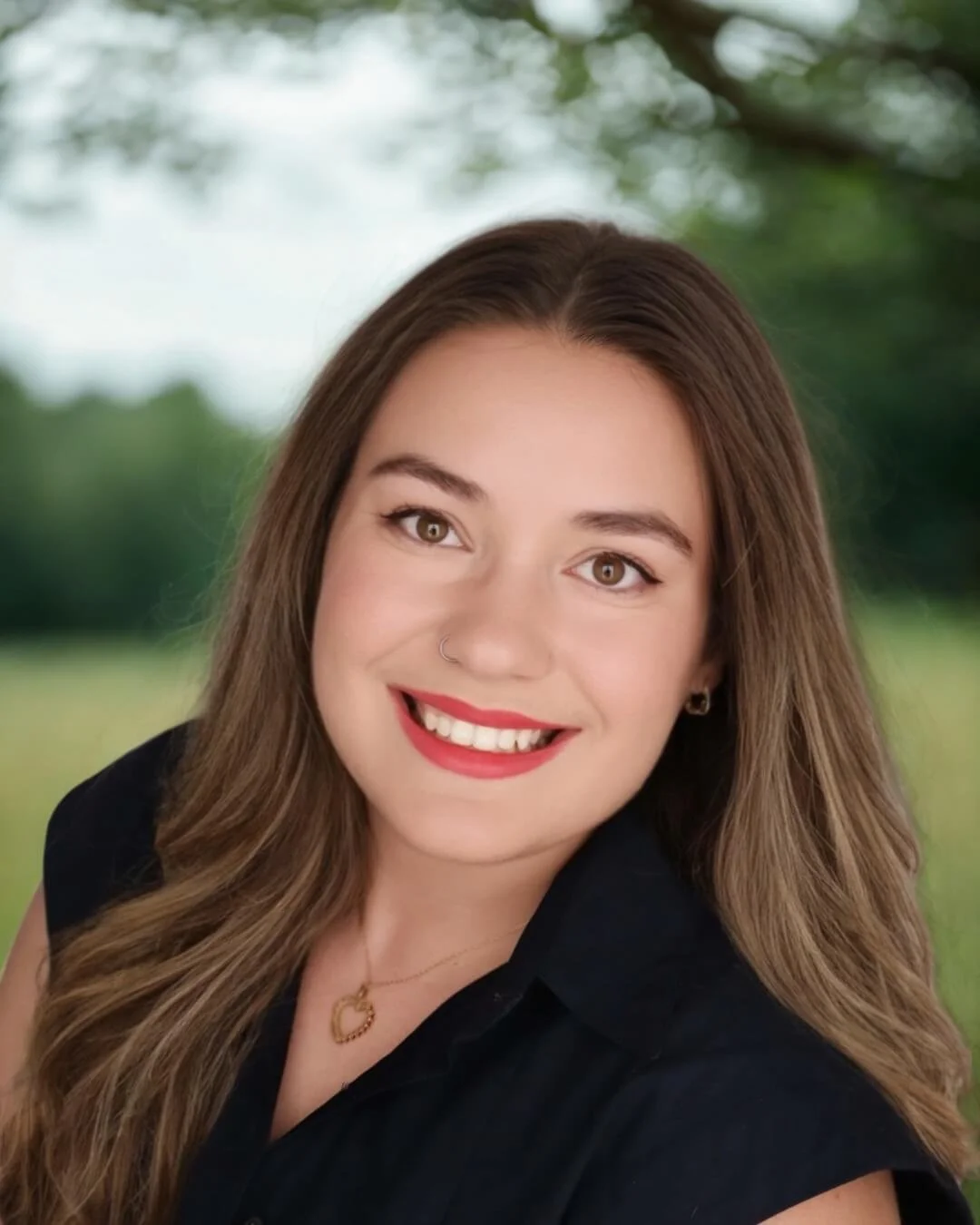 Kristen Johns Headshot: smiling young female therapist with long wavy brown hair, wearing a black top, gold necklace, and earrings, outdoors with blurred greenery in the background.