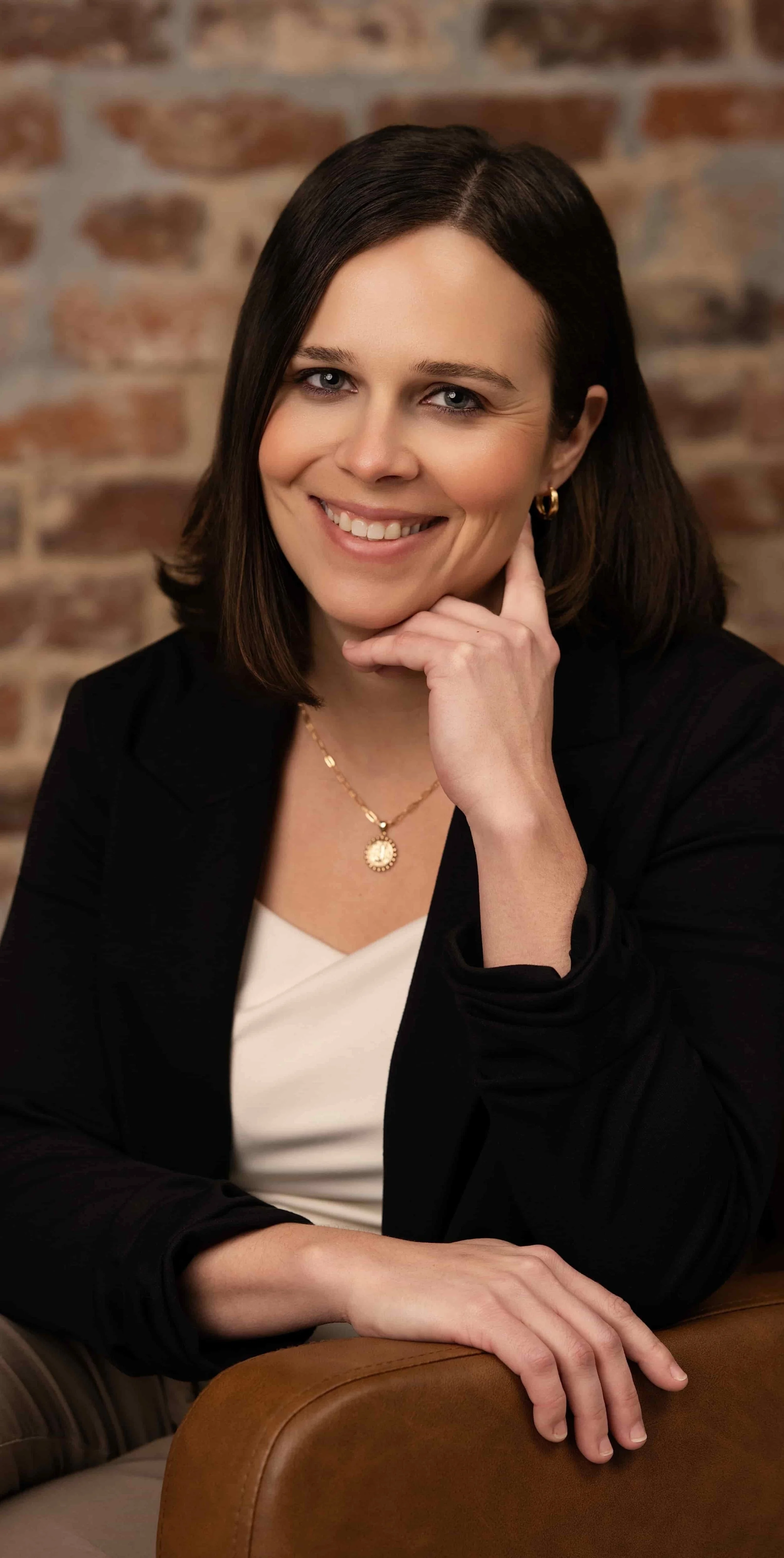 Professional headshot of a smiling female therapist seated in a brown leather chair, wearing a black blazer and light blouse, with a brick wall background.