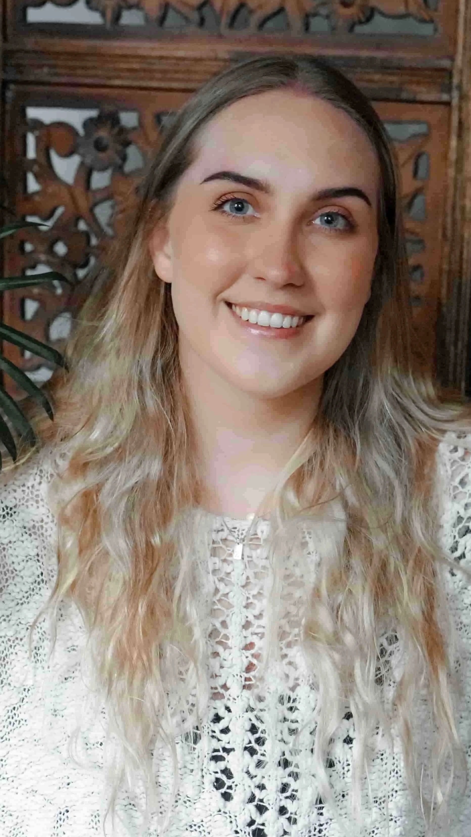 Taci Clay Headshot: A young female therapist with long, wavy blonde hair and blue eyes smiling at the camera, wearing a white lace blouse and is sitting in front of a wooden decorative background with a leafy green plant partially visible on her left
