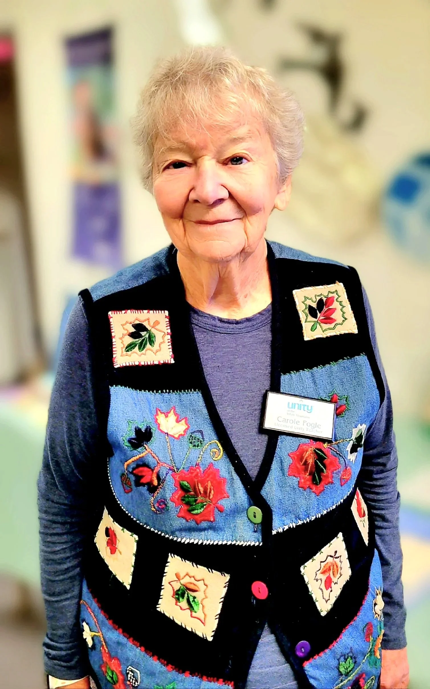 An elderly woman with short gray hair smiling, wearing a blue, black, and beige sweater with embroidered fall leaves and acorns, and a name tag that reads 'Carole Fogle, Licensed Unity Teacher.' The background is blurred with colorful posters and objects.